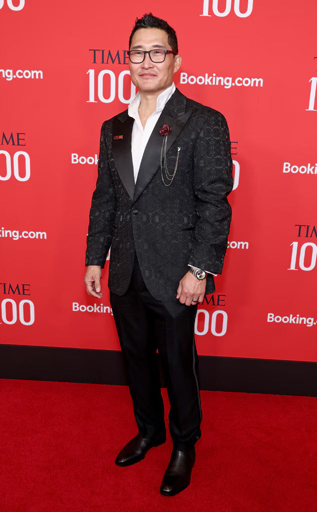 Man wearing a patterned black tuxedo jacket with satin lapels, white dress shirt, black trousers, and black shoes standing on a red carpet in front of a red backdrop with TIME 100 and Booking.com logos