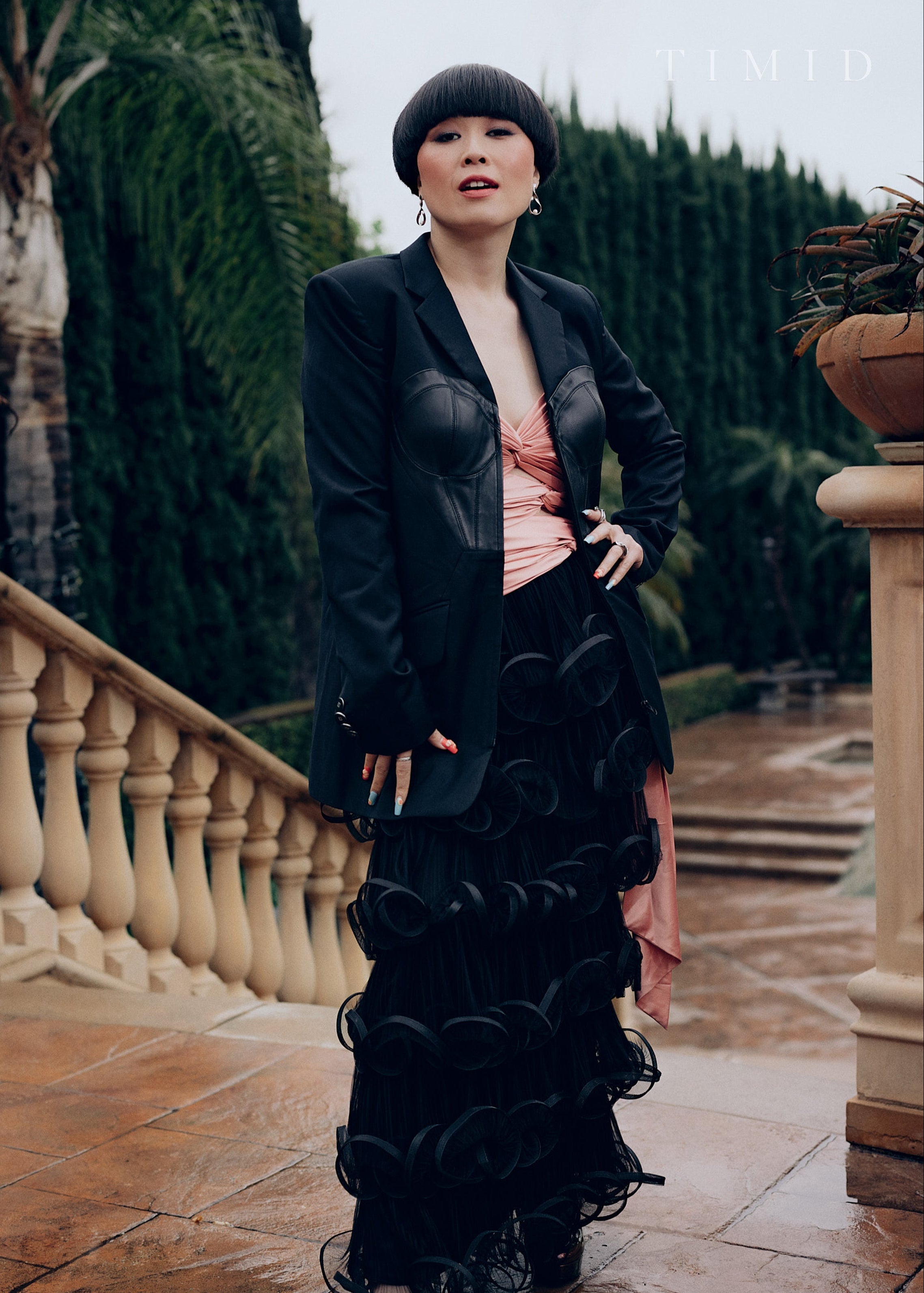 A woman poses outdoors near a stone balustrade wearing a tailored black blazer over a structured black and pink dress, with lush greenery in the background.