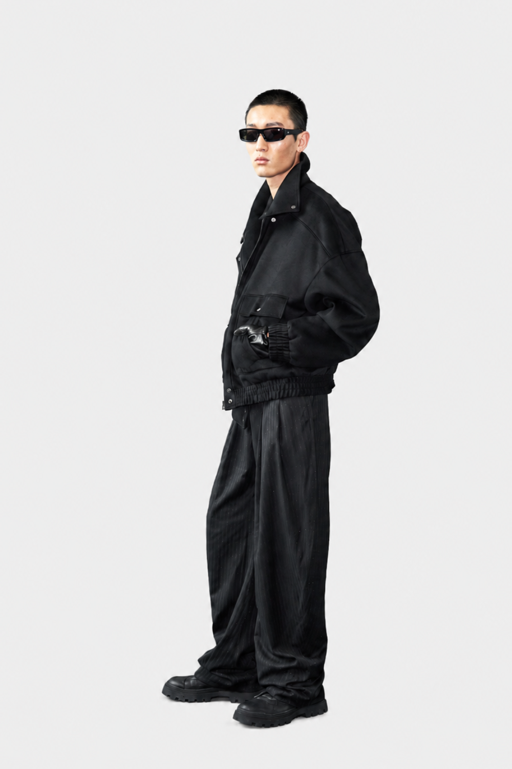 A model wearing a black oversized jacket, matching black wide-leg pants, black sunglasses, and black sneakers, standing in profile against a light gray studio background.