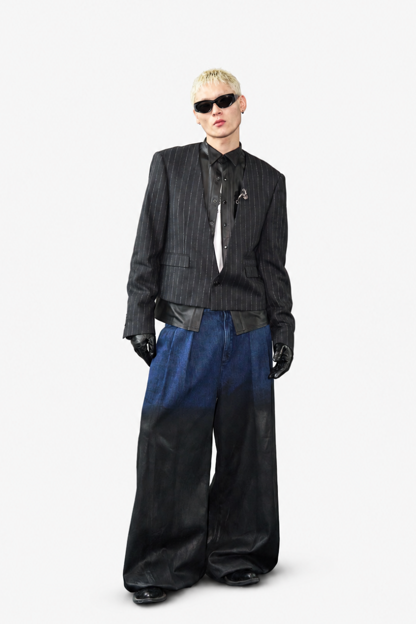 A model with short blonde hair and sunglasses wearing a black pinstripe blazer, black shirt, blue-to-black ombre wide-leg trousers, and black gloves, standing against a light gray background.