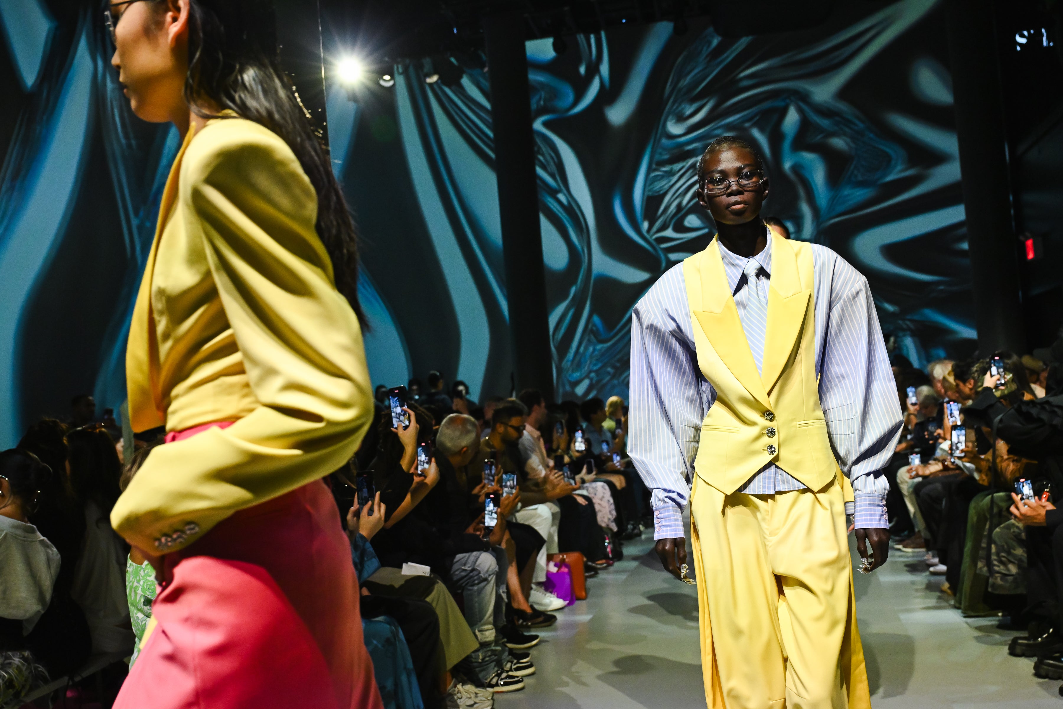A model walks down a runway wearing a yellow tailored vest with wide lapels over a blue and white striped shirt featuring voluminous sleeves, paired with matching yellow trousers against an abstract, wavy backdrop.