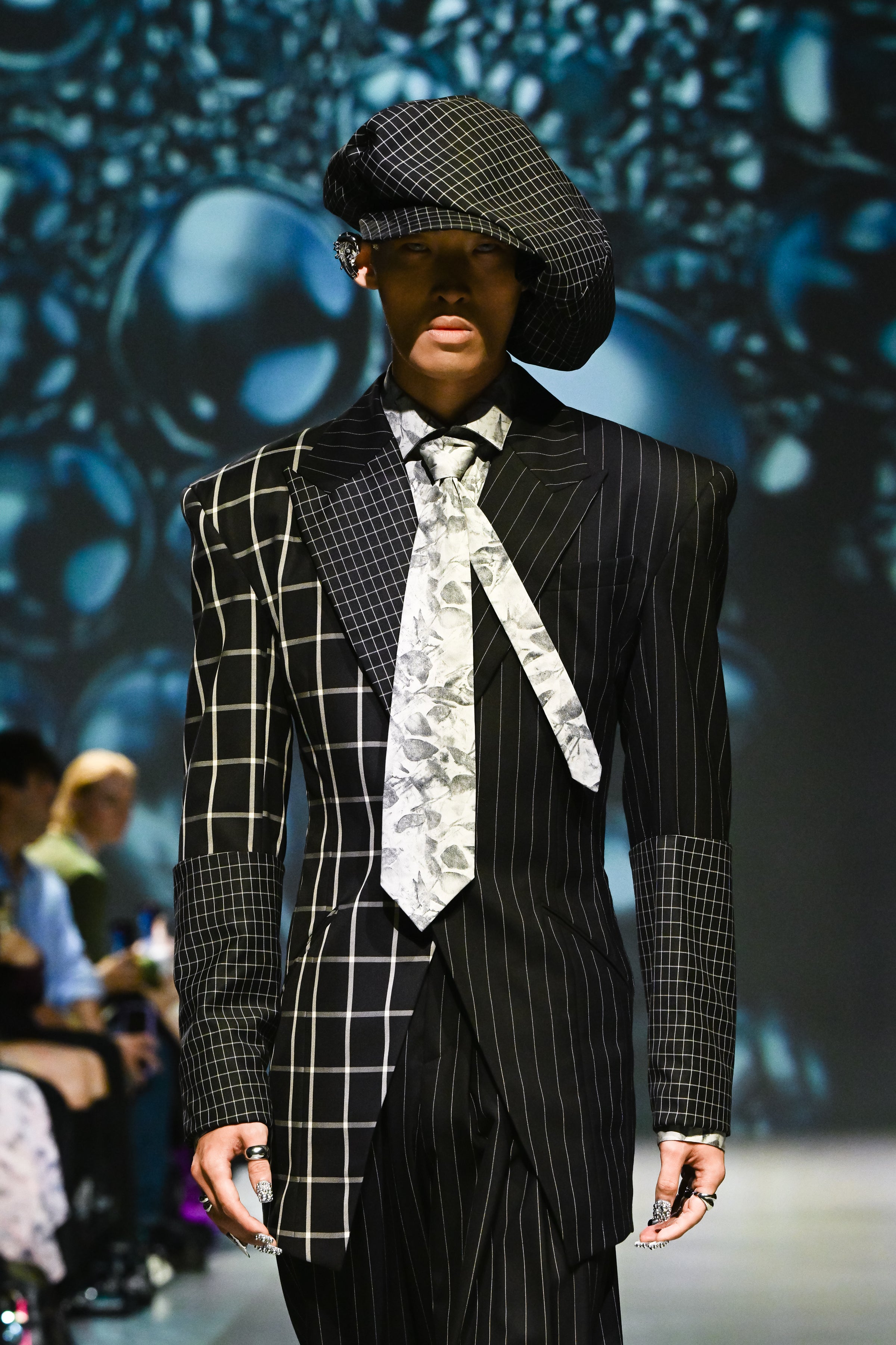 Model on runway wearing a mixed-pattern black blazer with pinstripe, windowpane, and check panels, paired with Black Wide-Leg Trousers showcasing bespoke tailoring and a sculptural silhouette.