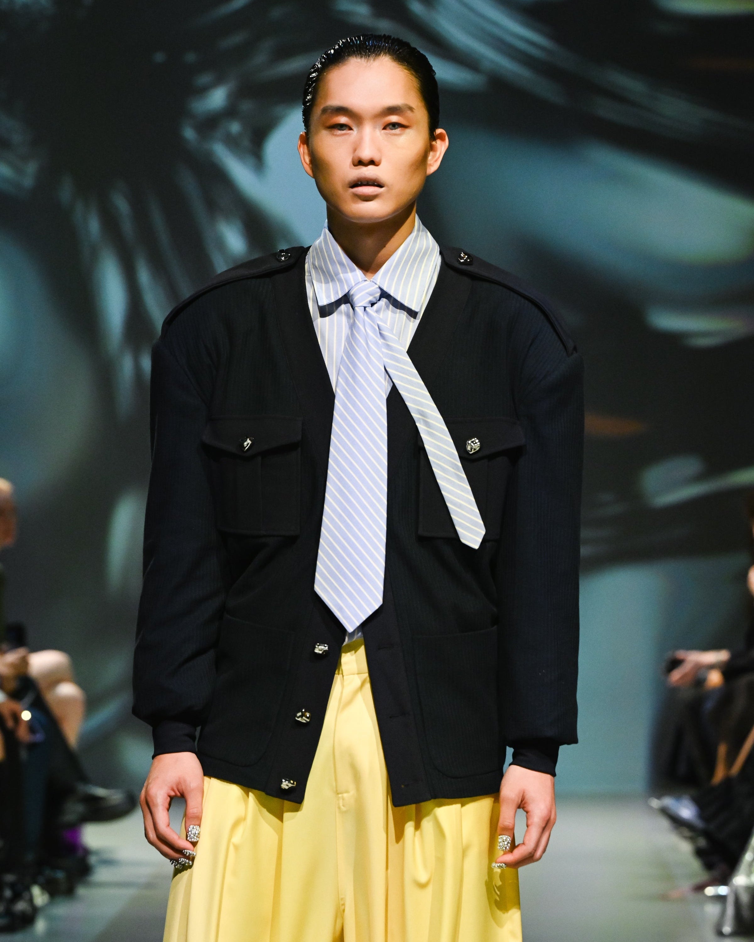 Runway model wearing a Navy Military-Style Utility Cardigan with structured flap pockets, epaulettes, and decorative buttons, styled with a loosely knotted striped tie and wide-leg yellow trousers for a sculptural silhouette in luxury designer fashion.