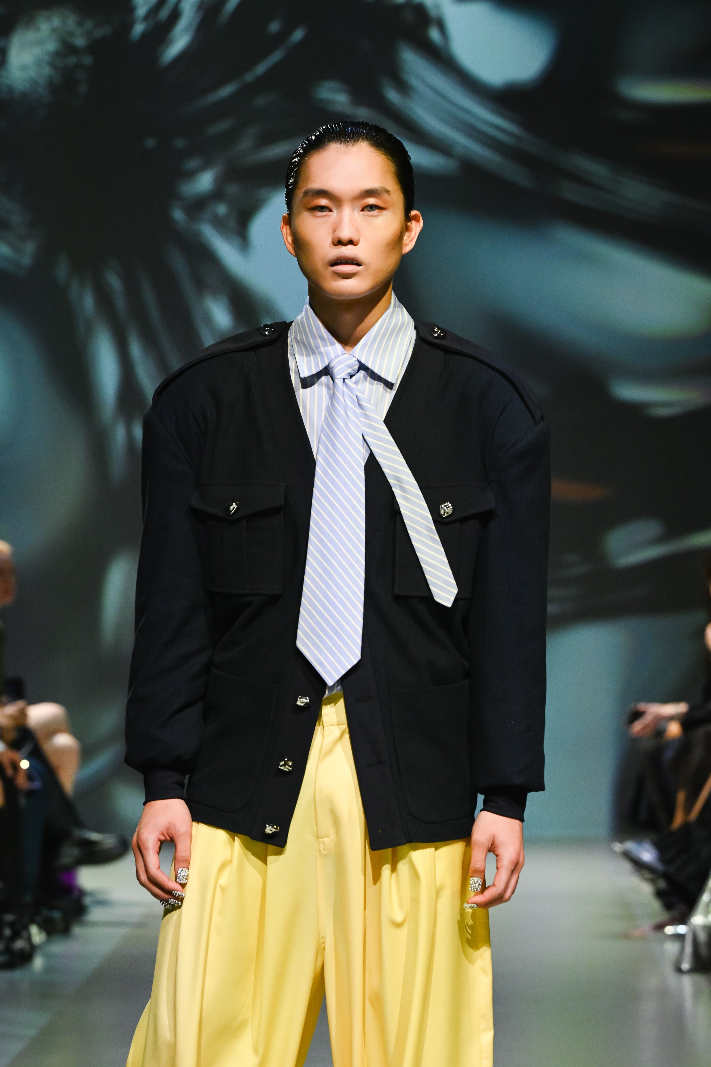 Runway model wearing a Navy Military-Style Utility Cardigan with structured flap pockets, epaulettes, and decorative buttons, styled with a loosely knotted striped tie and wide-leg yellow trousers for a sculptural silhouette in luxury designer fashion.