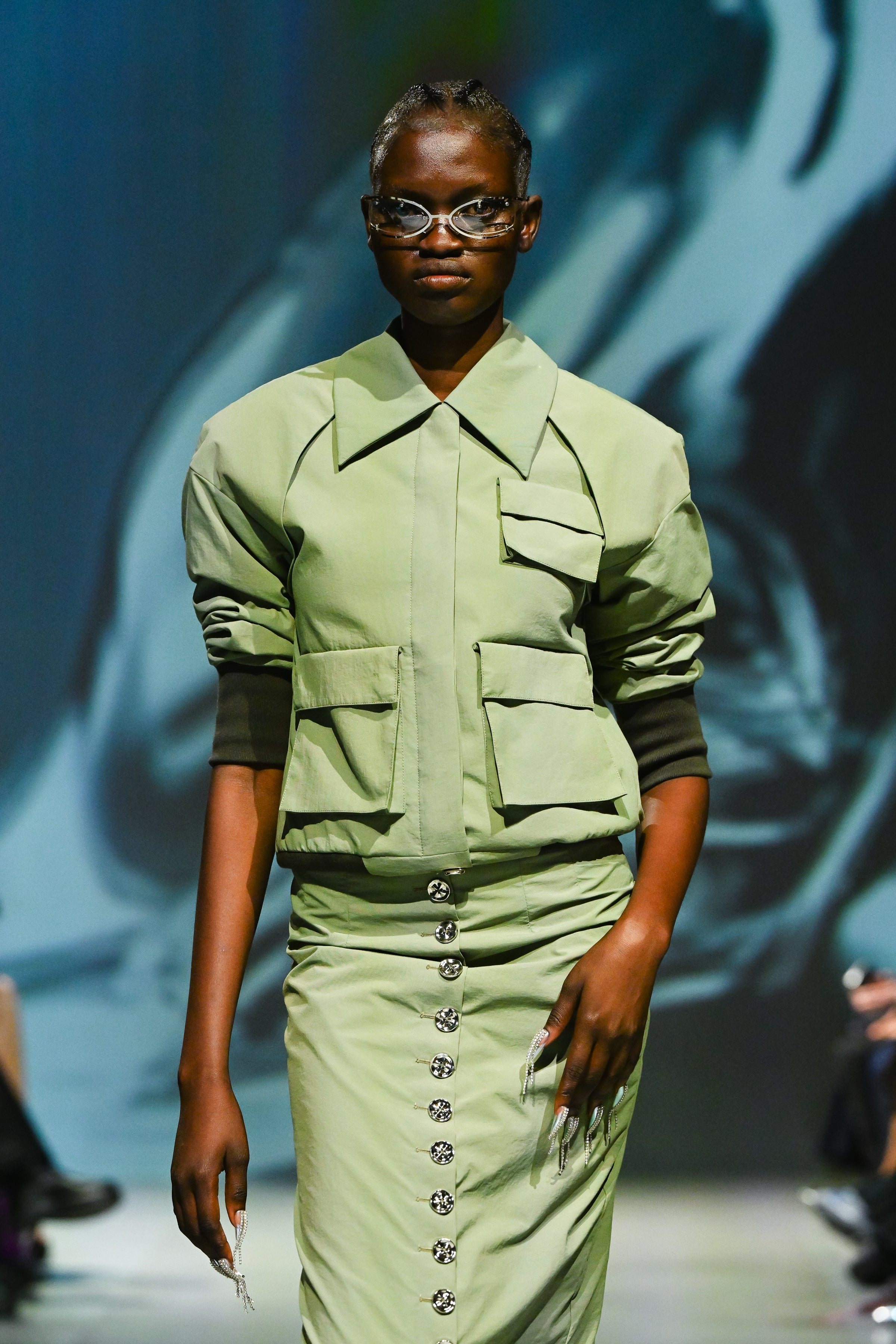Model walking runway wearing the Sage Green Utility Bomber Jacket styled with a high-waisted buttoned skirt, featuring sharp tailoring and a modern sculptural silhouette from SHAO New York luxury designer fashion.