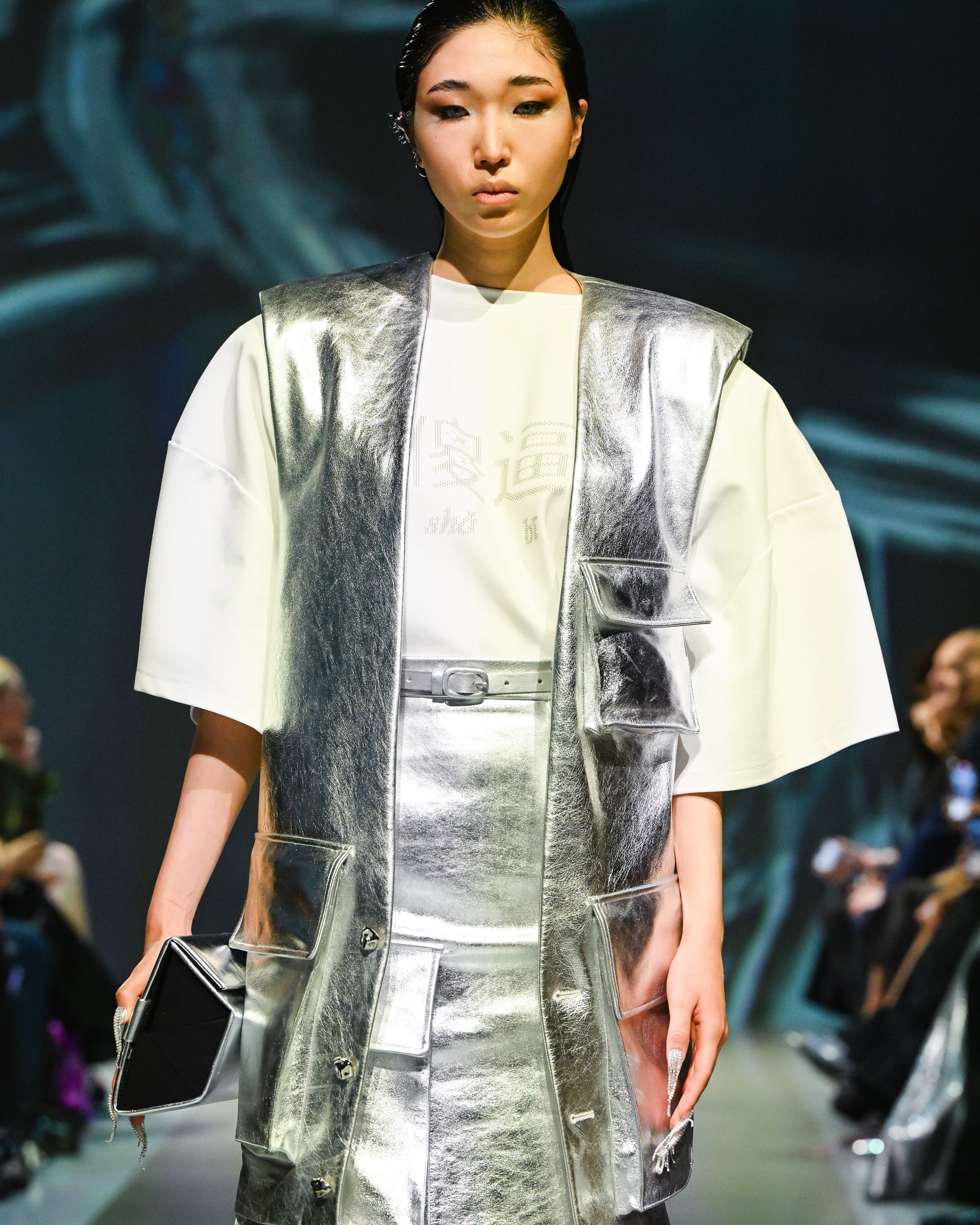 Model wearing a White Oversized Tee with Bold Chinese Graphics, styled with a sleeveless metallic silver vest and matching skirt, showcasing a futuristic sculptural silhouette in luxury designer fashion.