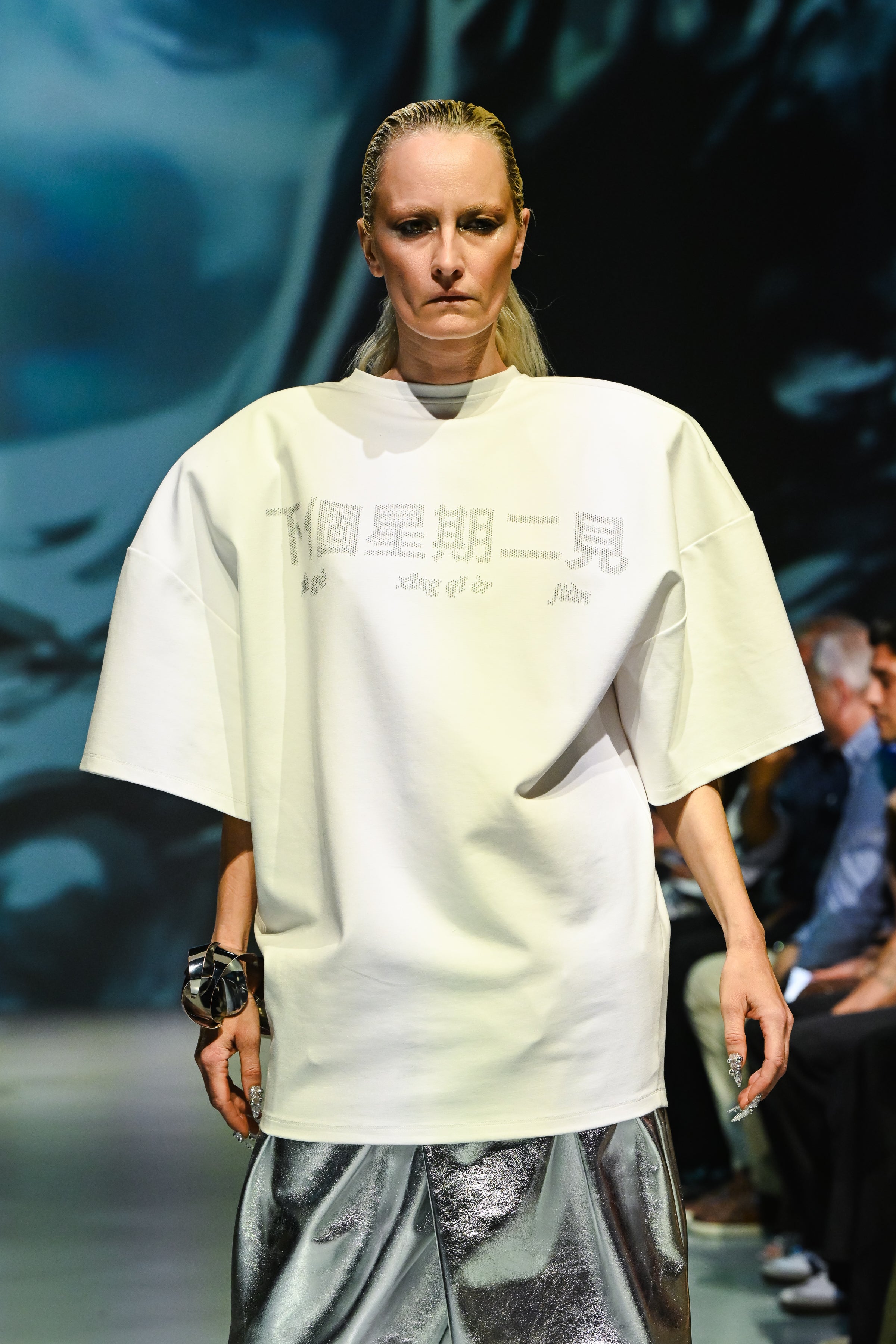 Model walking runway in White "See You Next Tuesday" Oversized Graphic Tee with Chinese Text, styled with metallic silver wide-leg pants and a bold black bracelet, representing SHAO New York luxury designer fashion.