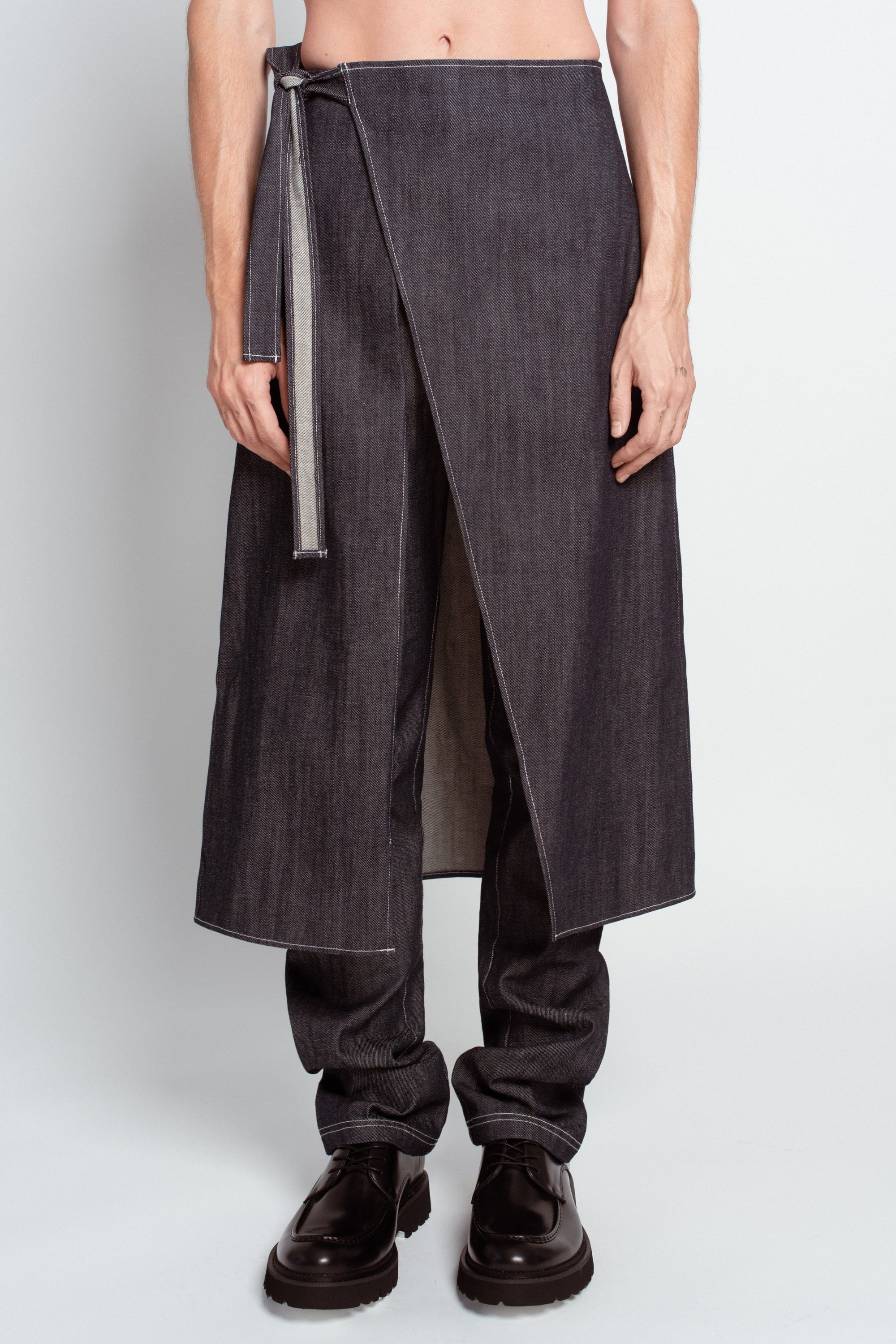 Front view of a model wearing Charcoal Denim Dual-Layer Trousers – Wrap Skirt Construction with a minimalist wrap skirt overlay and relaxed denim trousers underneath, paired with black formal shoes.