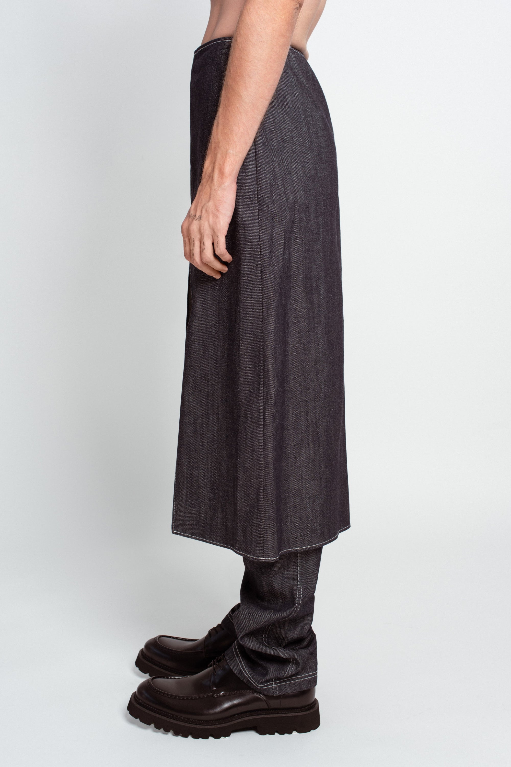 Side view of Charcoal Denim Dual-Layer Trousers – Wrap Skirt Construction featuring a sleek wrap skirt overlay over relaxed denim trousers paired with black chunky dress shoes in a minimalist setting.