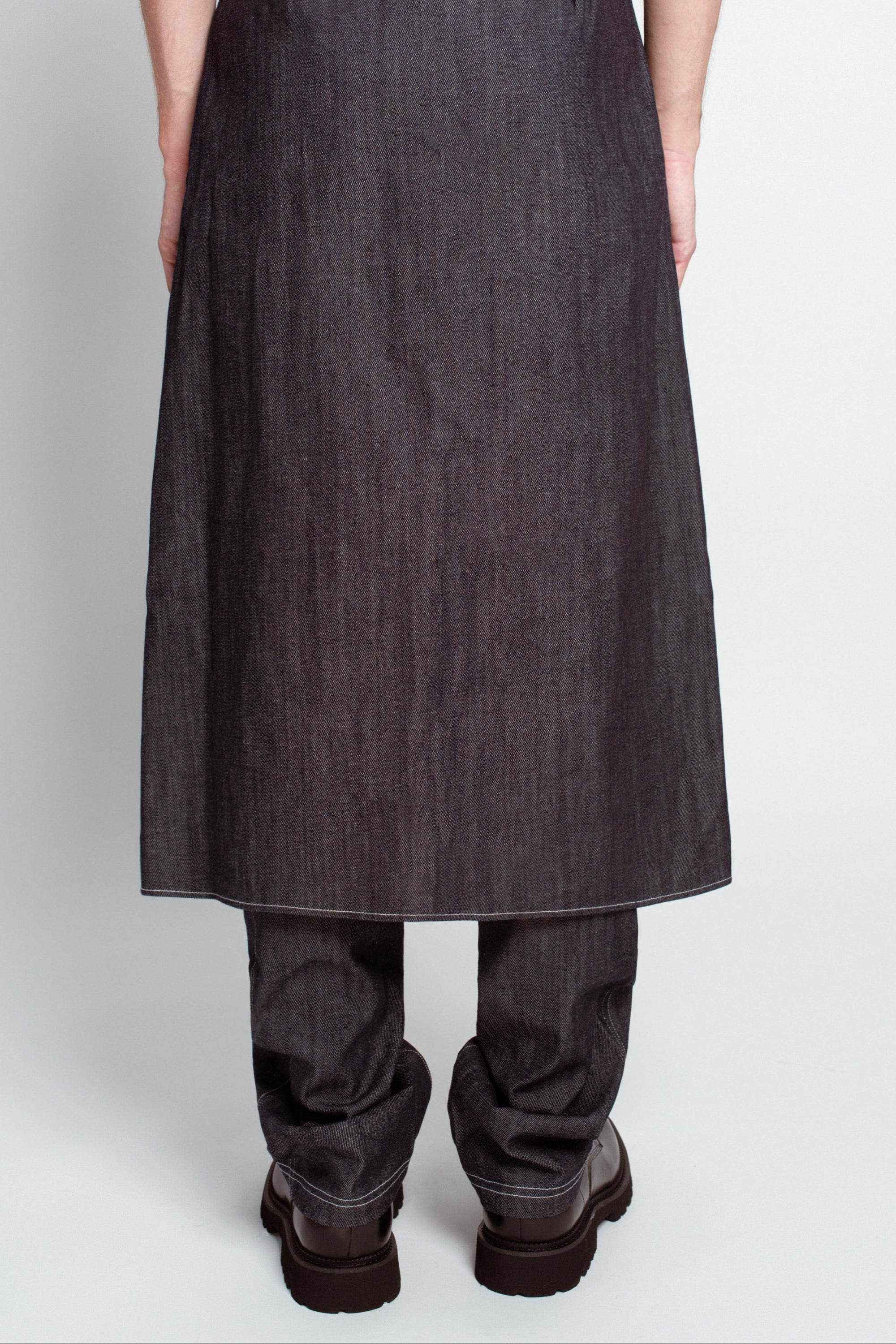 Back view of Charcoal Denim Dual-Layer Trousers – Wrap Skirt Construction featuring a long wrap skirt overlay in dark denim layered over relaxed trousers, paired with chunky black shoes for a sculptural luxury designer fashion look.