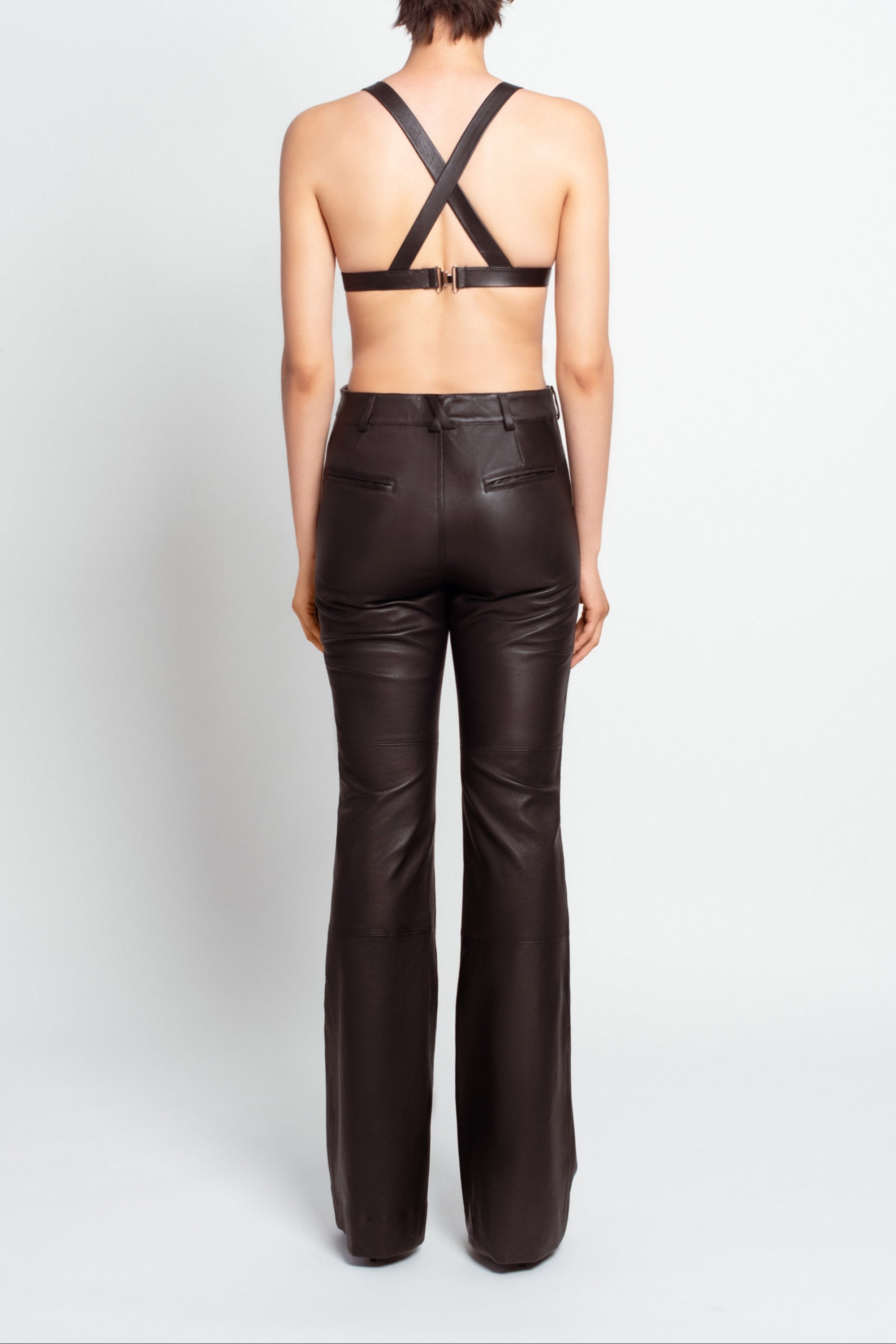 Back view of model wearing Jet Black Leather Bustier – Architectural Construction featuring crossed straps and buckle closure, paired with high-waisted black leather trousers for a sleek, sculptural silhouette.