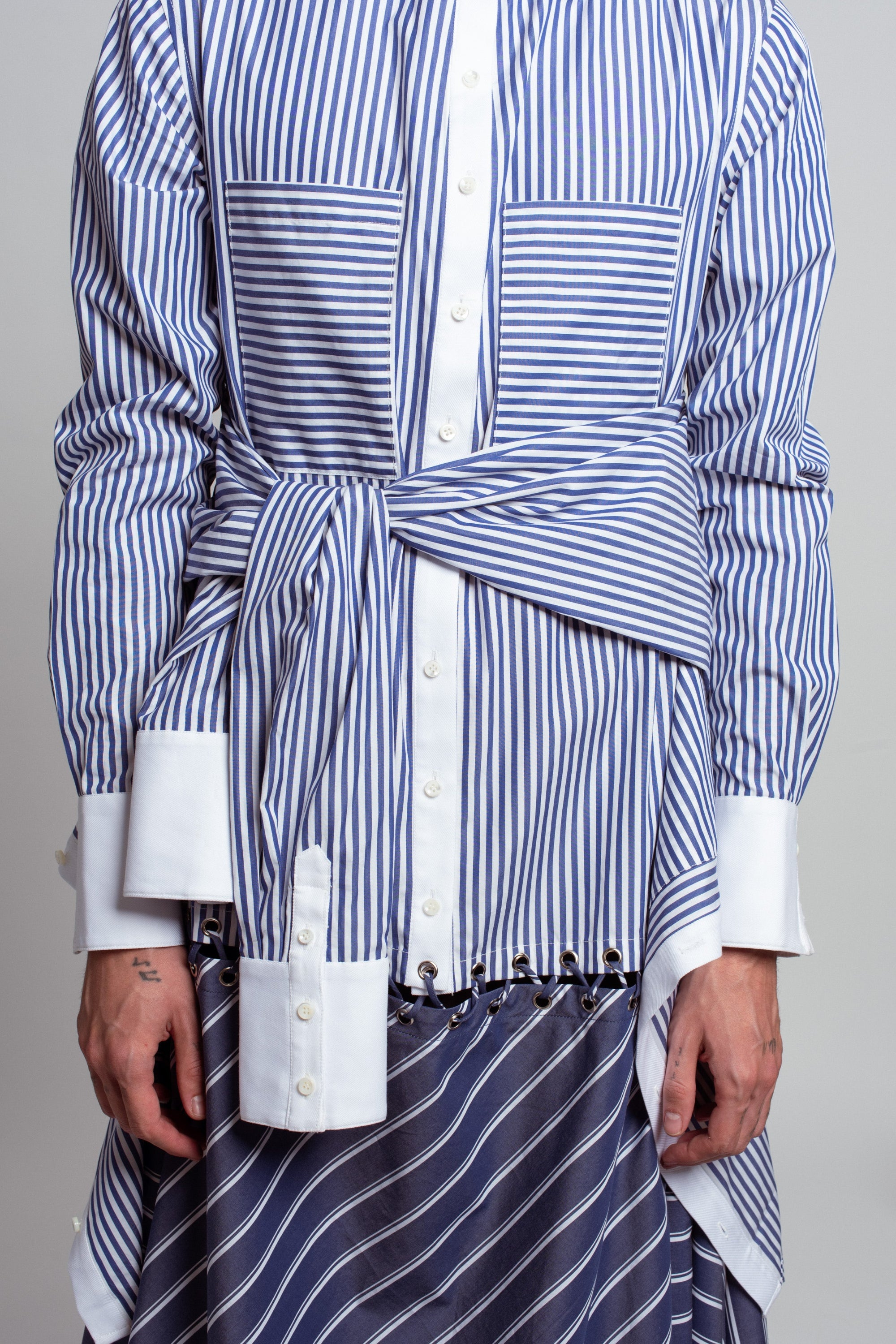 Close-up view of Blue Pinstripe Asymmetric Layered Shirtdress featuring contrasting horizontal and vertical stripes with oversized cuffs, a tied waist detail, and layered striped skirt, showcasing luxury designer fashion and a sculptural silhouette.