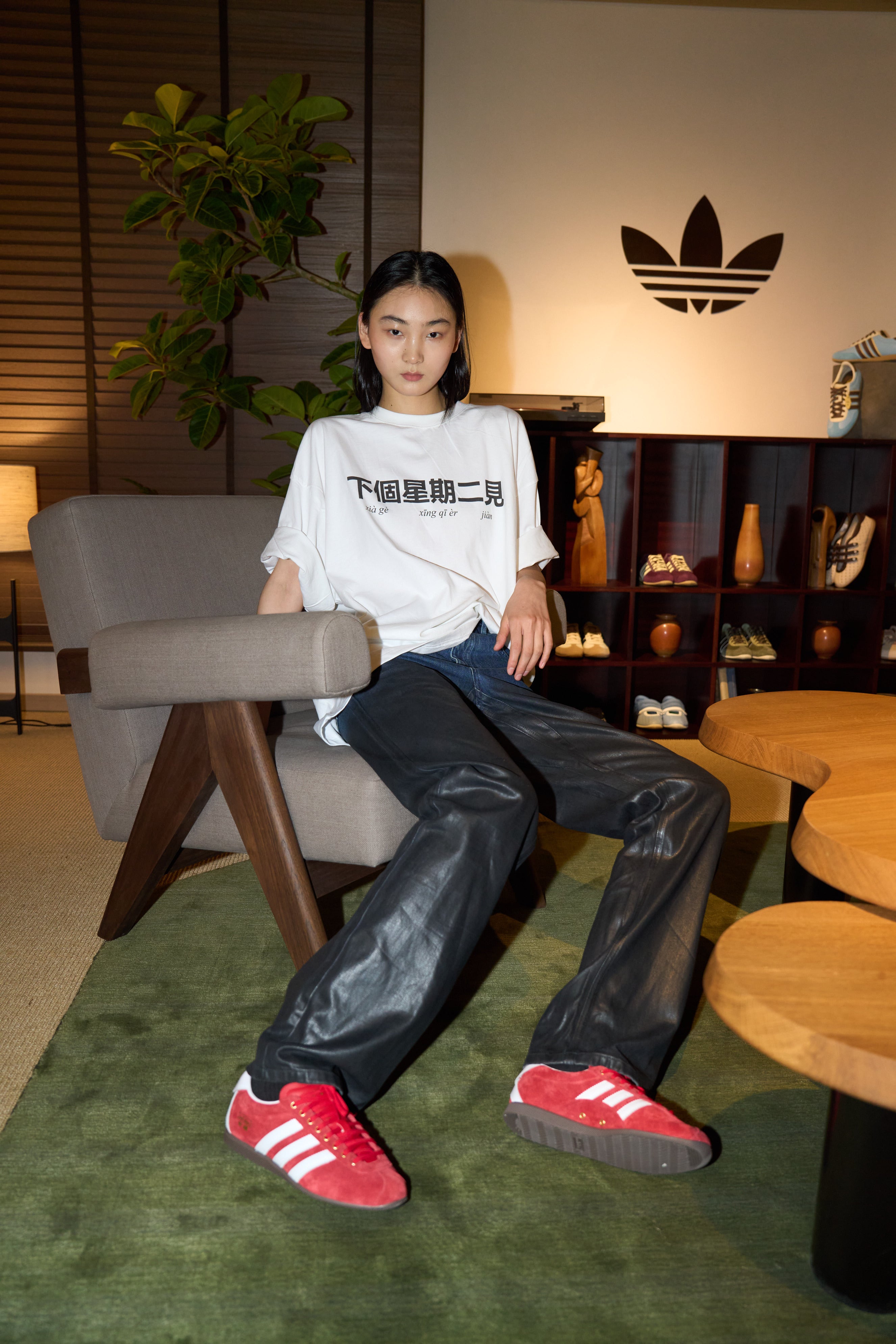 Model seated in a modern armchair wearing Drop-Crotch Denim Jeans with Leather Ombre Effect in a sleek black finish, paired with a relaxed white graphic T-shirt and red sneakers, embodying a casual yet sculptural silhouette in luxury designer fashion.