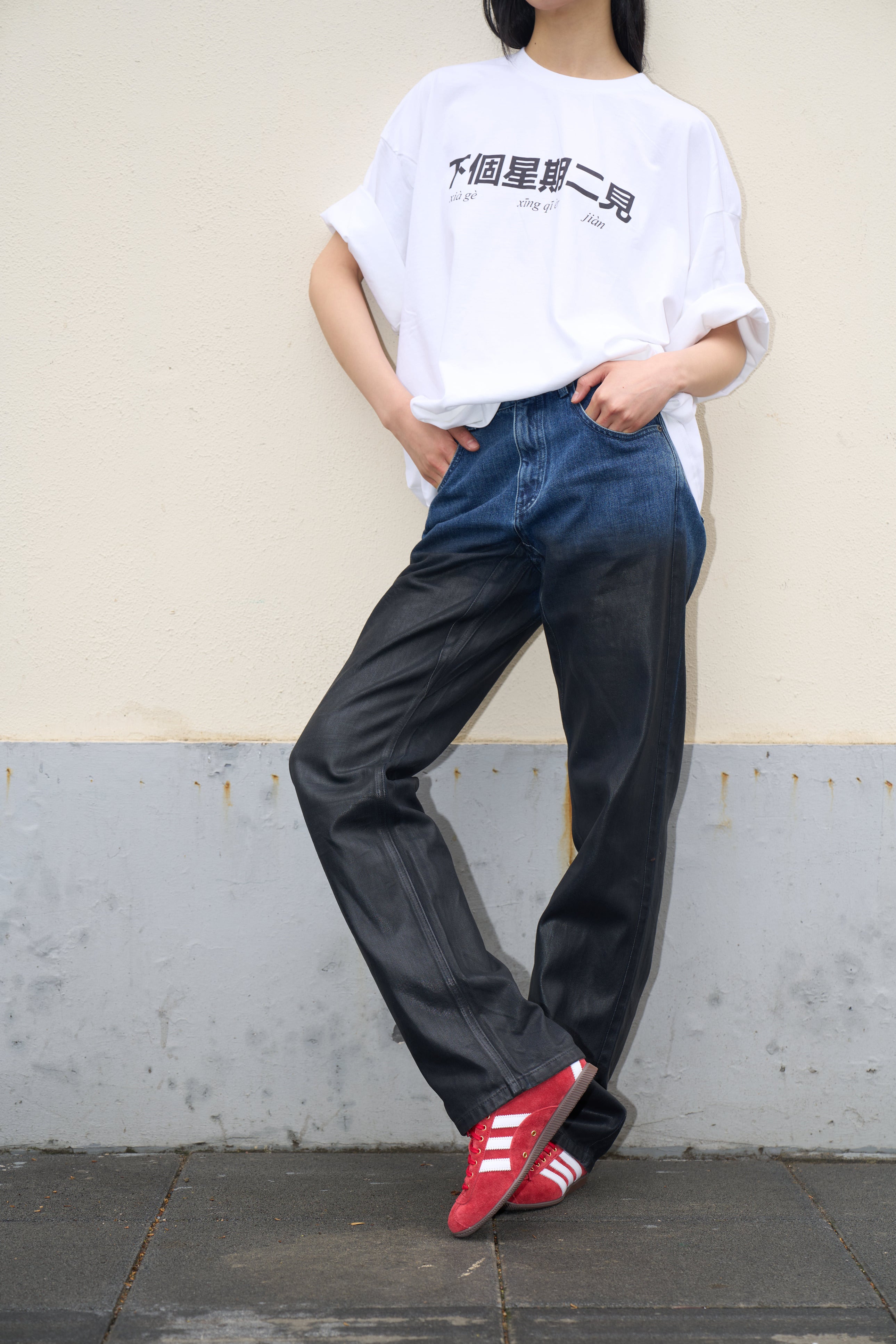 Model standing against a light wall wearing Drop-Crotch Denim Jeans with Leather Ombre Effect that transition from blue to black, paired with a loose white graphic T-shirt and bright red sneakers, presenting a laid-back yet refined sculptural silhouette in luxury designer fashion.