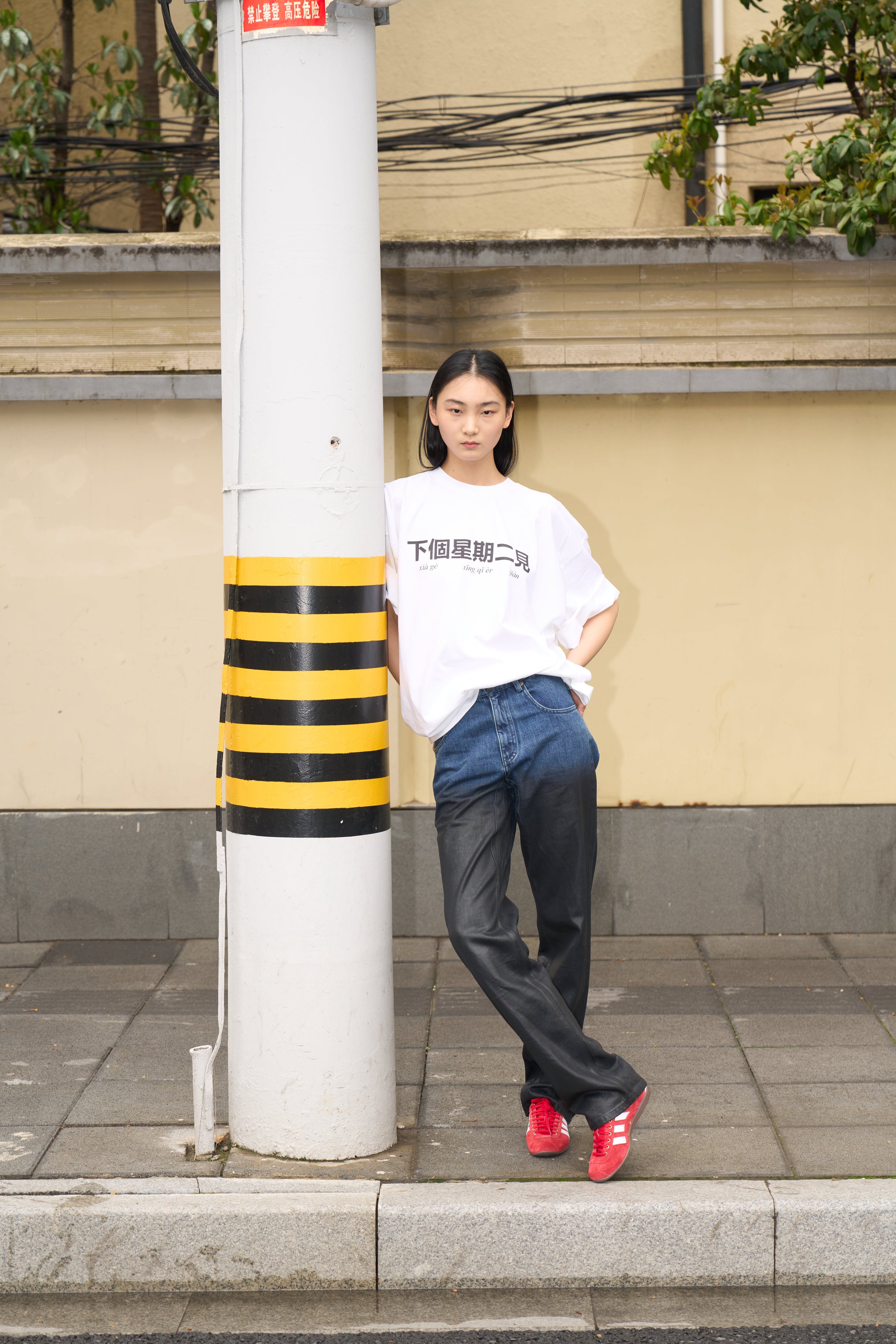 Young woman leaning against a striped utility pole wearing Drop-Crotch Denim Jeans with Leather Ombre Effect transitioning from deep blue to black, paired with a loose white graphic T-shirt and vibrant red sneakers, embodying a casual yet sculptural silhouette in SHAO New York luxury designer fashion.