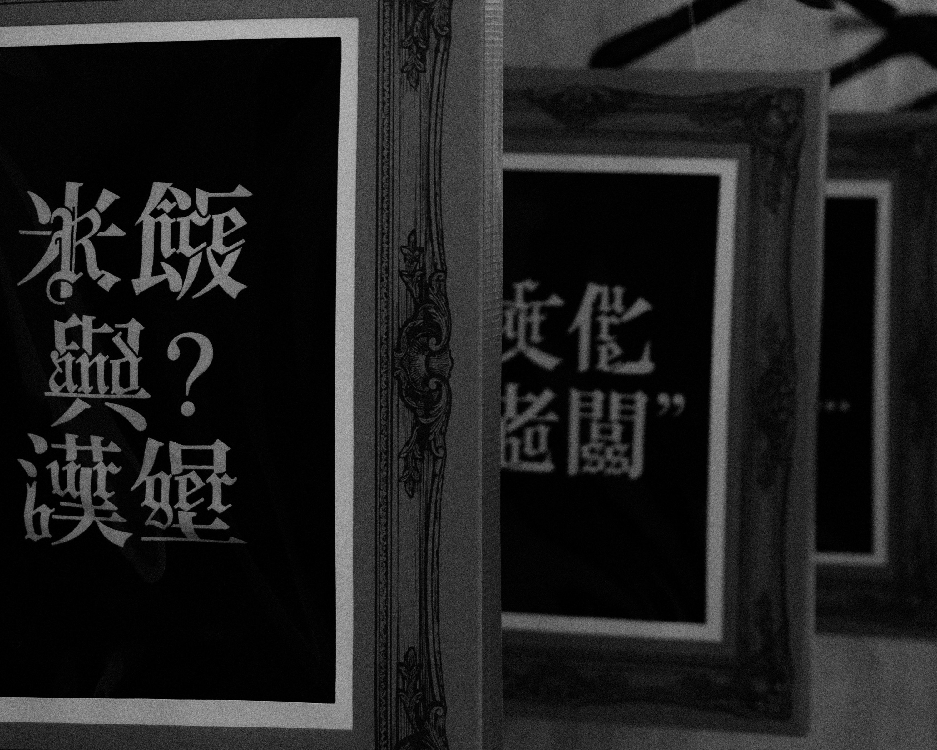 Black and white photograph of multiple ornate picture frames arranged in a row, each containing bold white Asian calligraphy characters on a dark background, creating a layered visual effect.