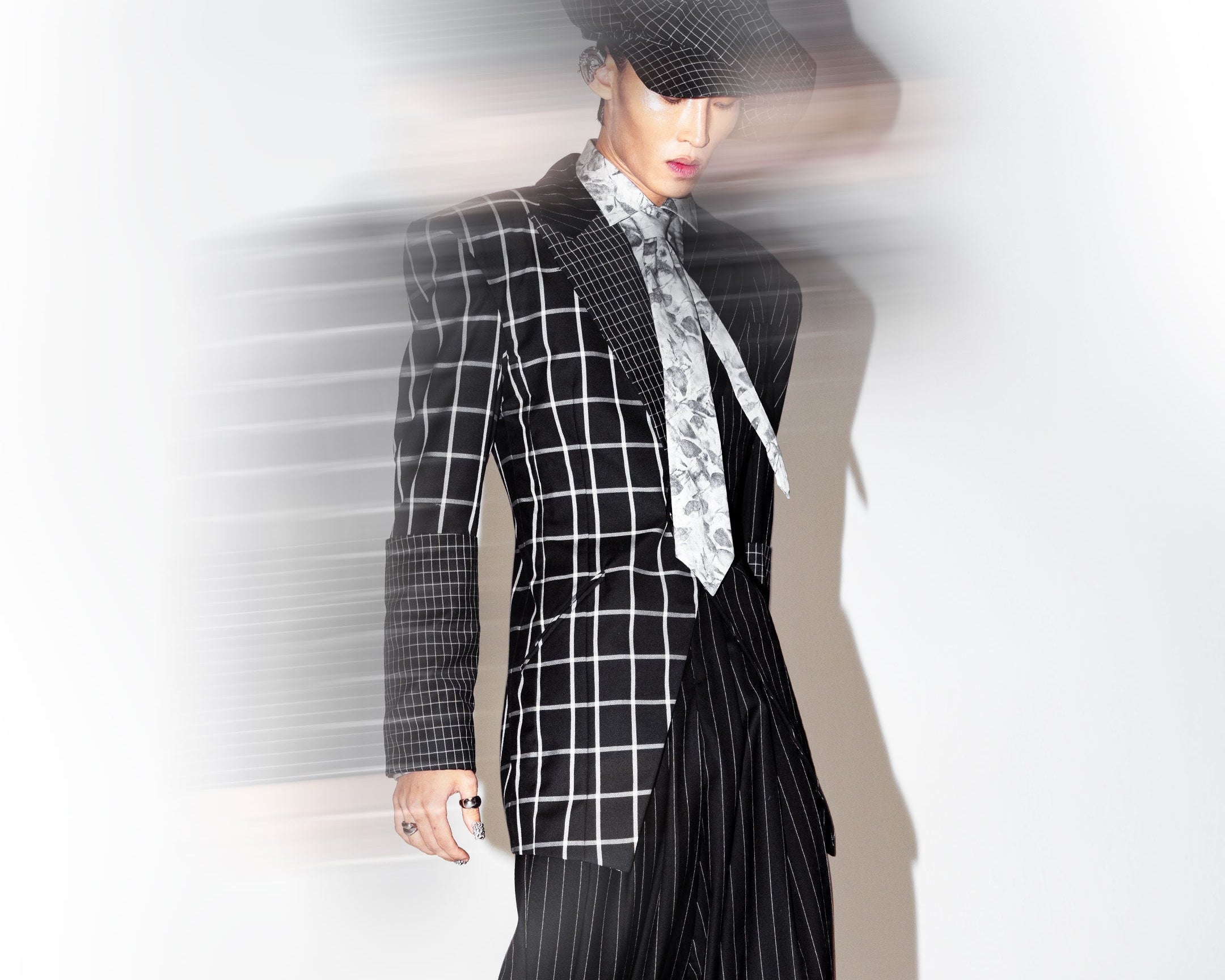 Model wearing a black and white checkered blazer with contrasting pinstripe sleeves and trousers, paired with a patterned tie and a black hat, captured with a motion blur effect.