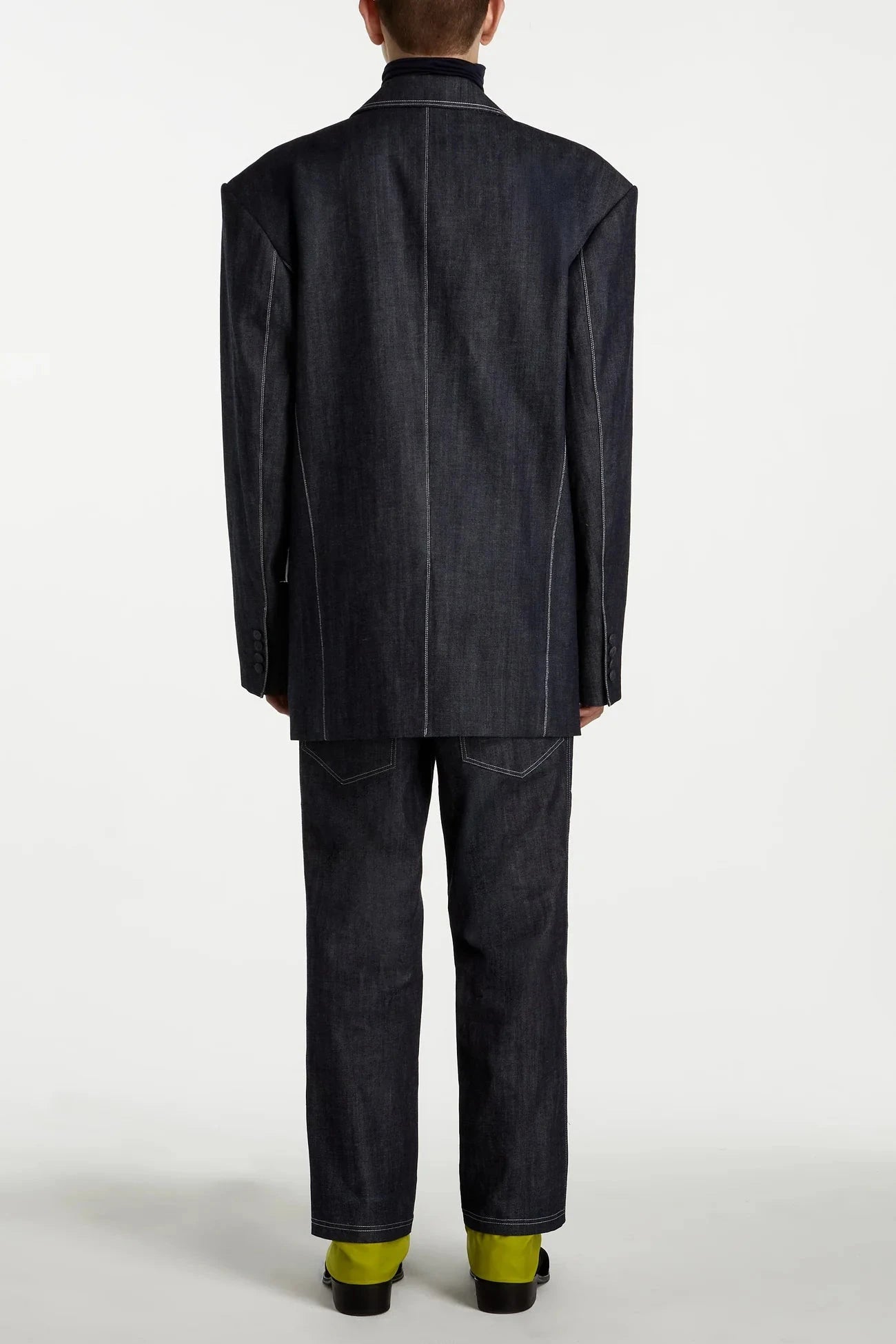 Model shown from the back wearing Dark Indigo Denim Blazer – Oversized Architectural Silhouette with pronounced vertical seams, paired with matching denim trousers and lime green shoes in a minimalist studio setting.