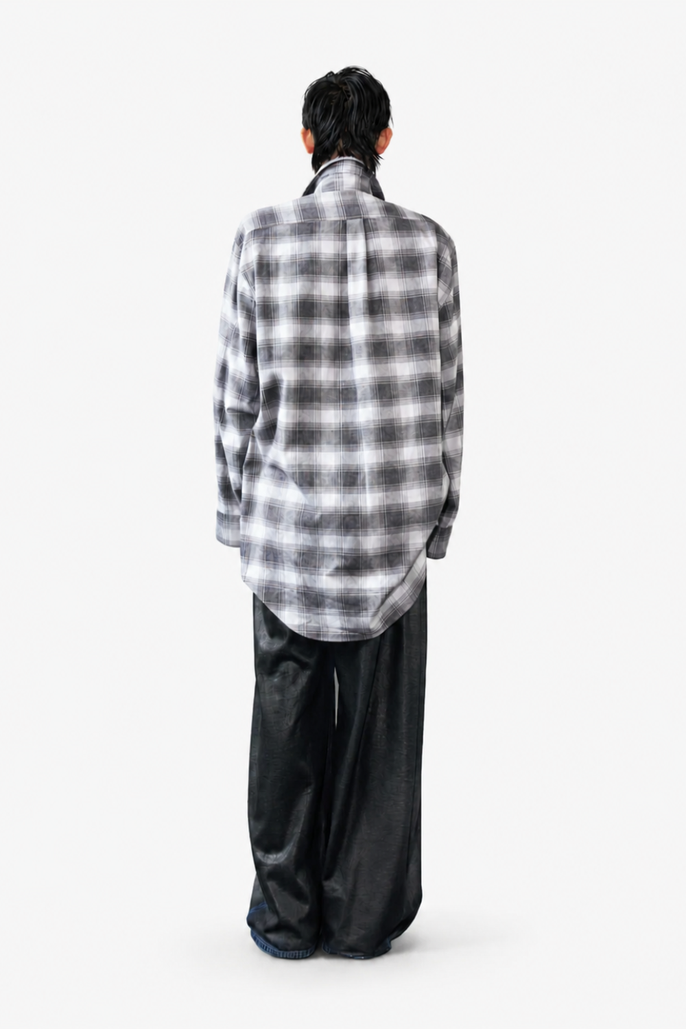 Model seen from behind wearing Box-Pleated All Over Coated Denim Jeans in a dark finish paired with a loose, long-sleeve grey plaid shirt, illustrating a relaxed sculptural silhouette in SHAO New York luxury designer fashion.