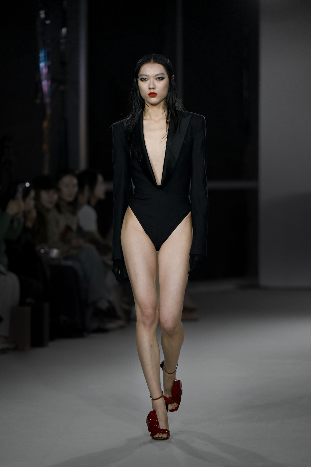 Model walking runway in the Tuxedo Bodysuit featuring a deep plunging neckline, tailored long sleeves with subtle satin lapels, creating a sculptural silhouette paired with red high-heeled sandals.
