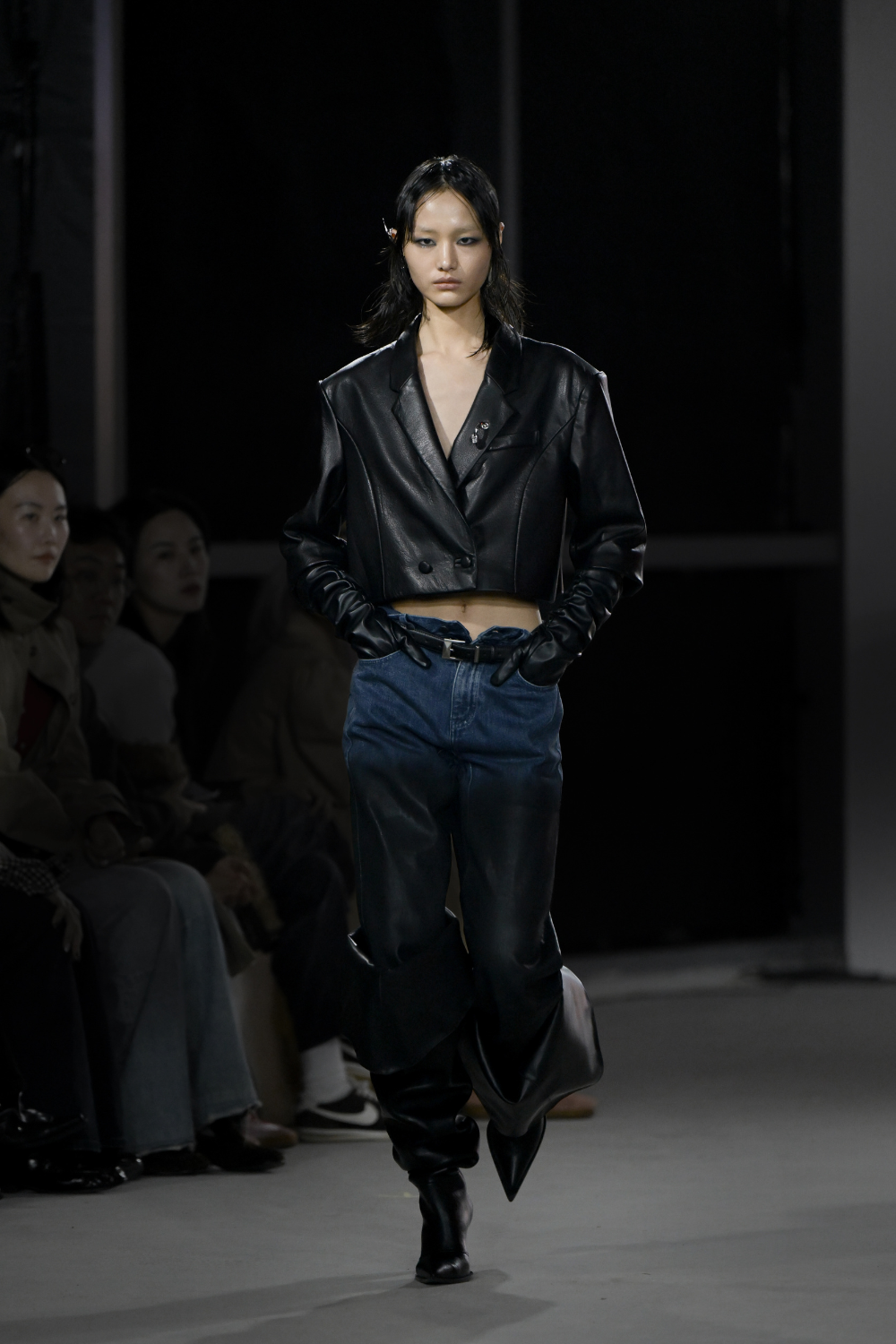 Model wearing Cropped Leather Double-Button Blazer with Piercing Detail in black, paired with high-waisted two-tone denim trousers, long leather gloves, and pointed leather boots, showcasing a sharp sculptural silhouette and bespoke New York designer tailoring.