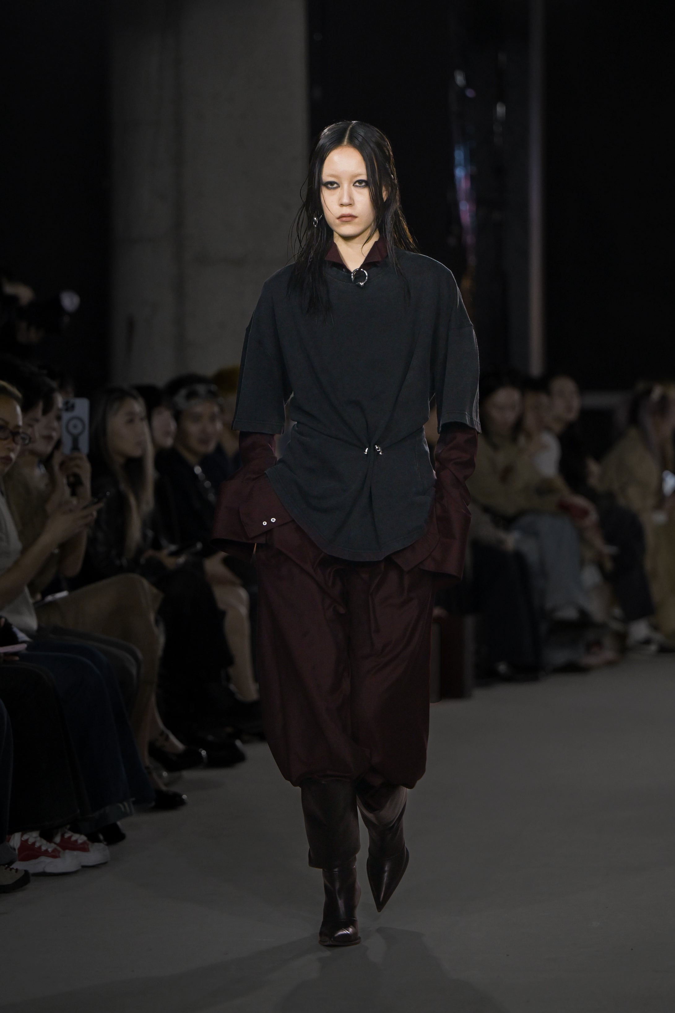 Model walking runway wearing Wool Barrel Pleated Trousers - Burgundy paired with layered dark tops featuring structured draping and asymmetrical styling, completed with black pointed boots, reflecting SHAO New York’s luxury bespoke tailoring and sculptural silhouette.