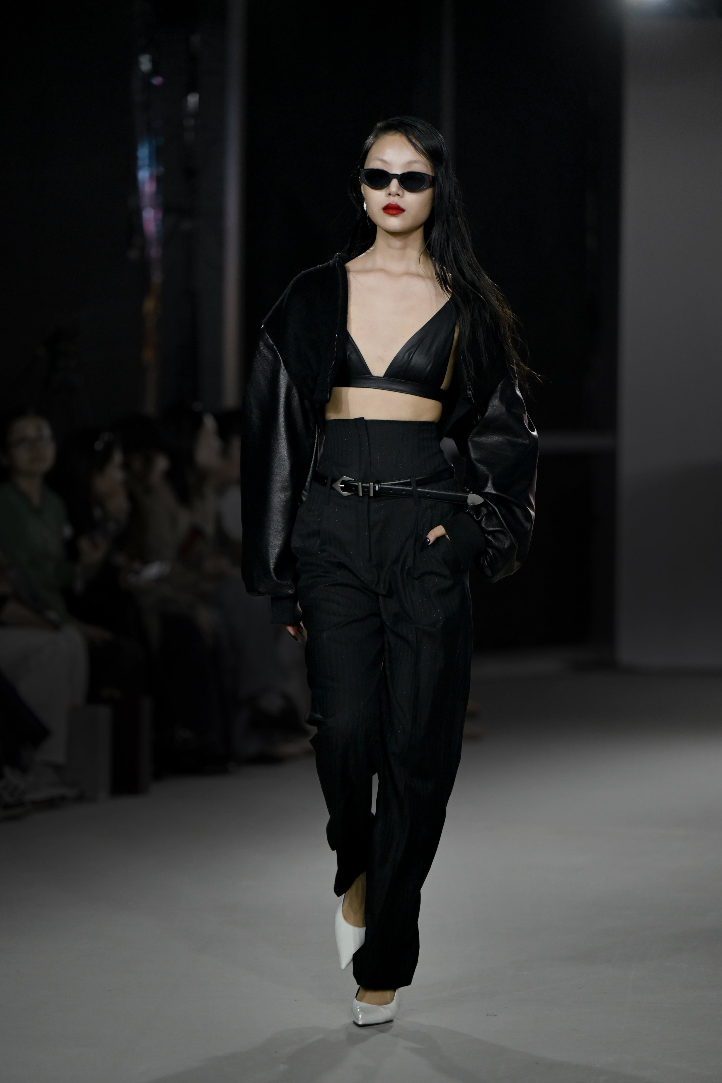 Model walking runway wearing the Cropped Alpaca and Leather Bomber Jacket styled with a black leather bralette, high-waisted pinstriped trousers, black belt, white pointed heels, and dark sunglasses, embodying SHAO New York’s luxury designer fashion with a sleek sculptural silhouette.