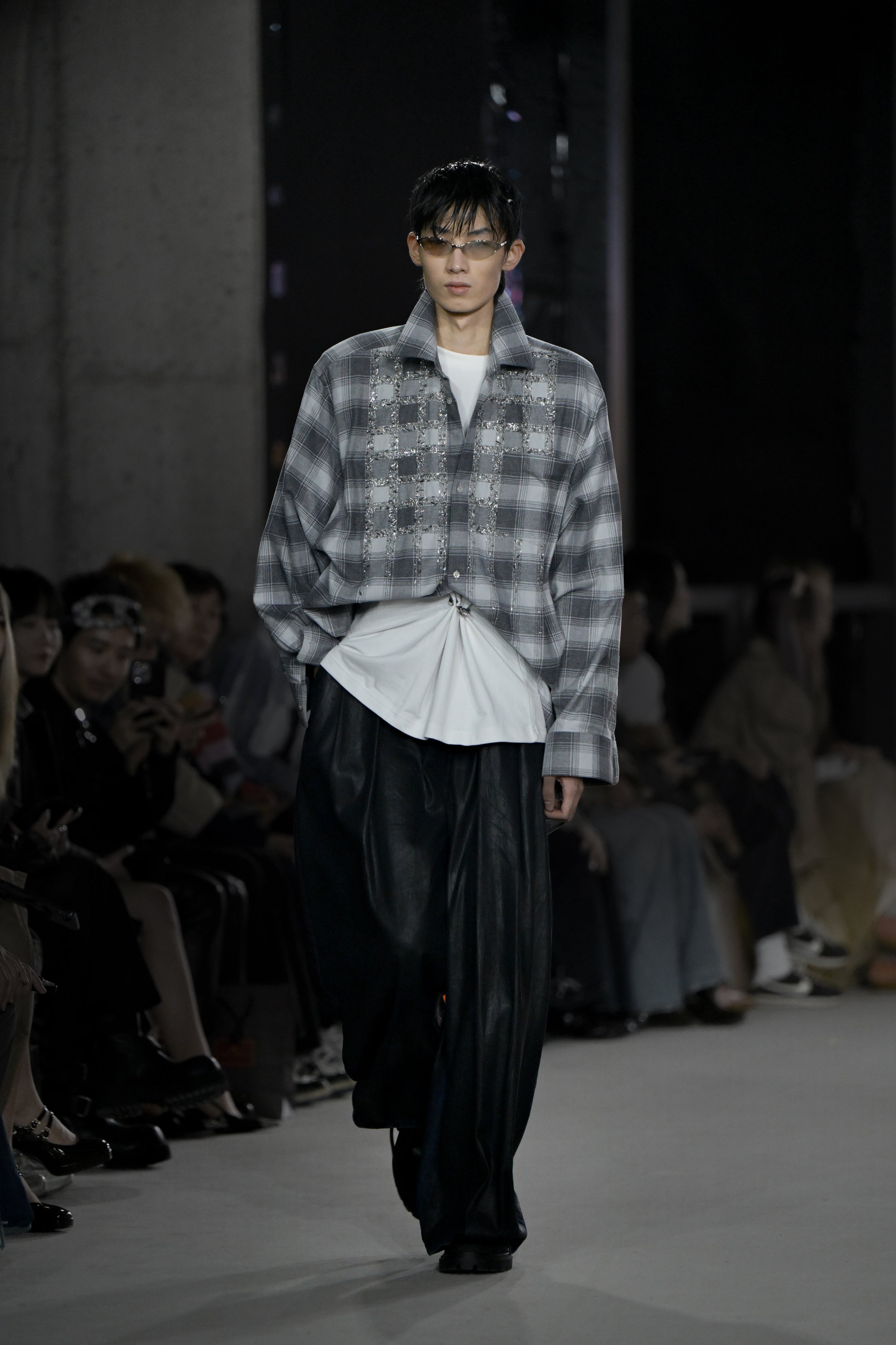 Model walking runway wearing the Pierced Vintage T-Shirt - White styled underneath a grey plaid oversized jacket, paired with loose black trousers and sleek glasses for a modern layered look.