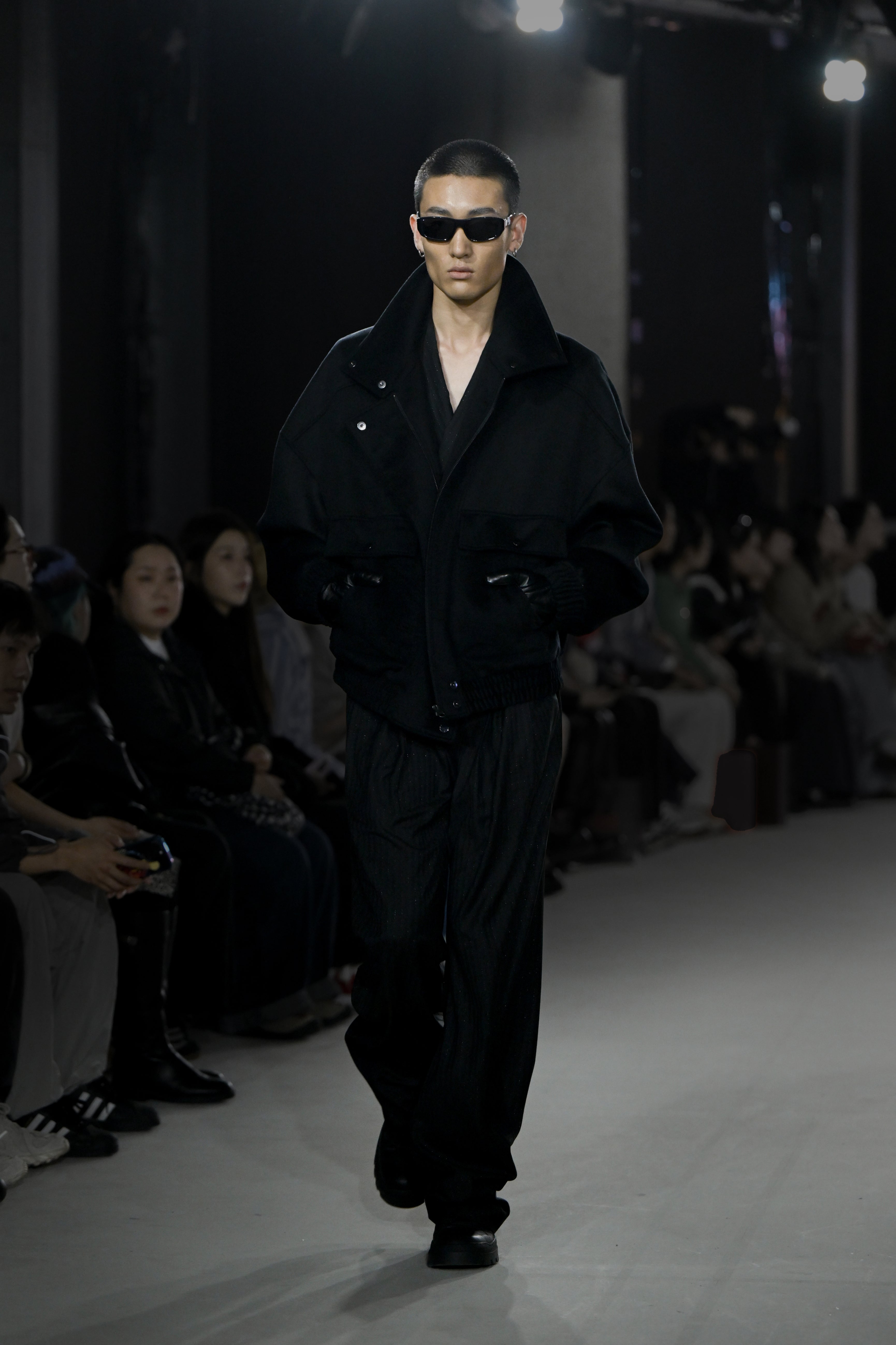 A model walks the runway wearing an oversized black jacket with a high collar and voluminous sleeves over loose black trousers, styled with black gloves and sleek sunglasses, showcasing a bold tailored look.