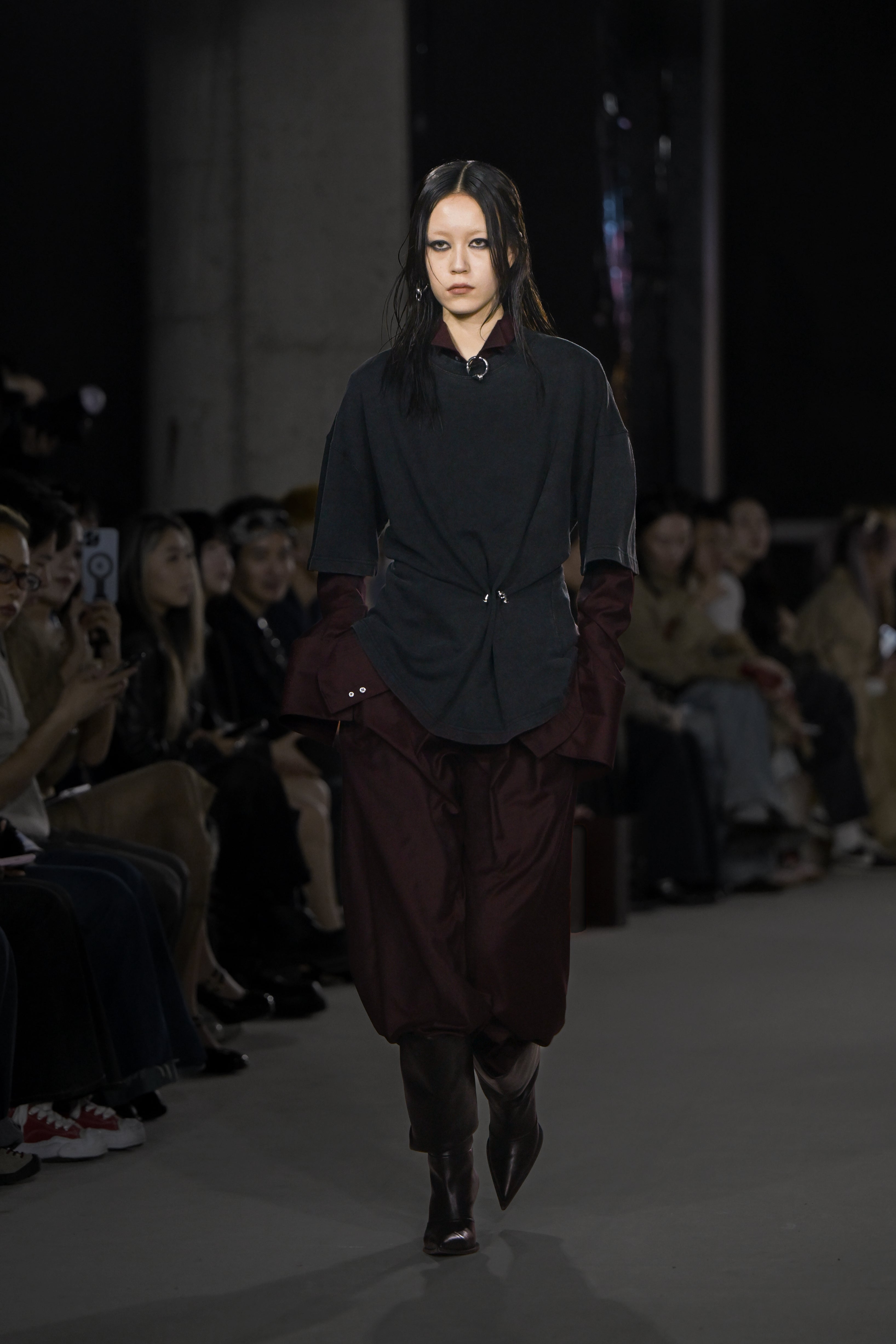 A model walks the runway wearing a layered ensemble featuring a loose black top cinched at the waist over a deep burgundy long-sleeve shirt and matching wide-leg pants, paired with dark pointed boots.