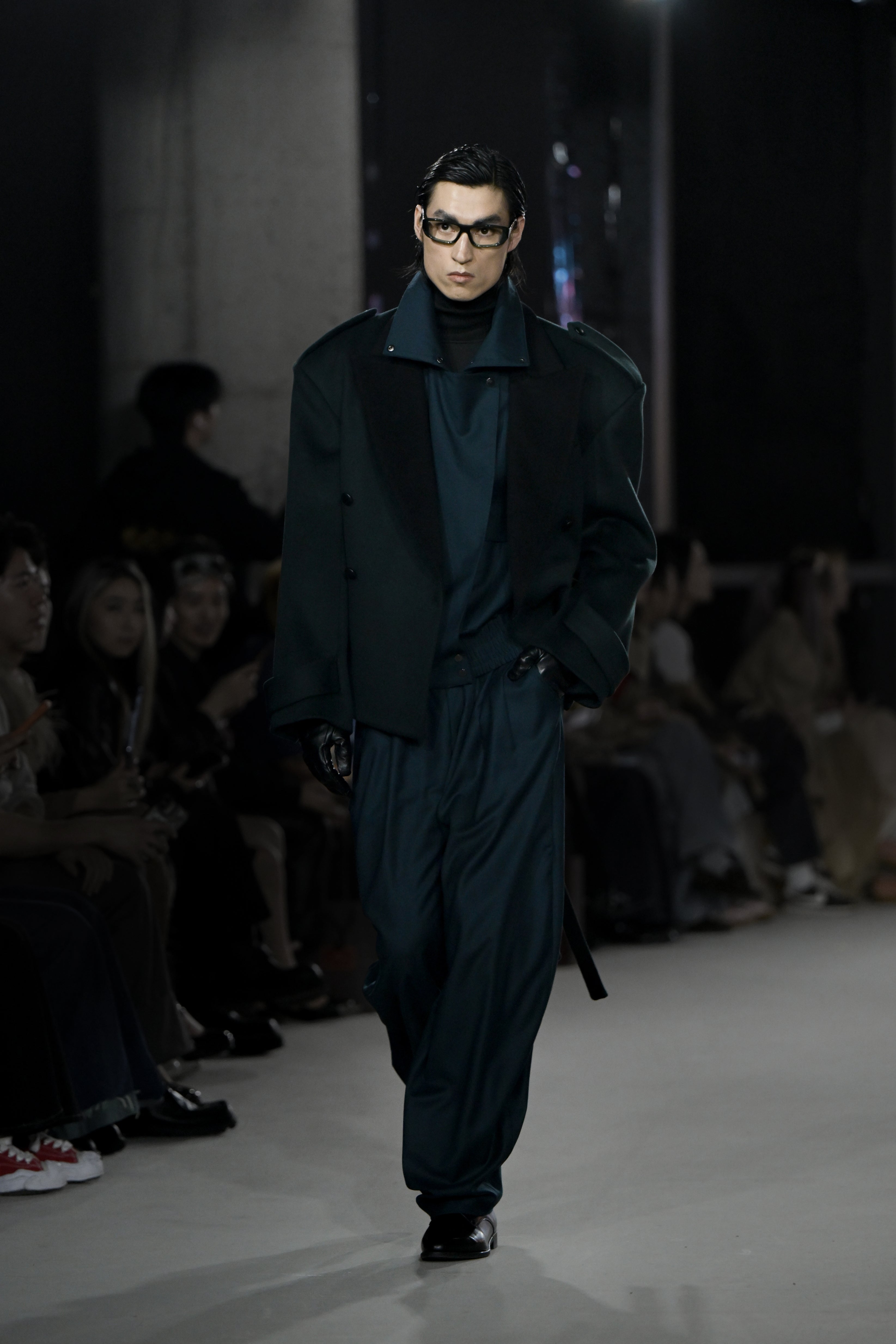 A model walks the runway wearing a dark oversized tailored jacket over a loose-fitting navy ensemble, styled with black gloves and statement glasses for a refined sculptural silhouette.
