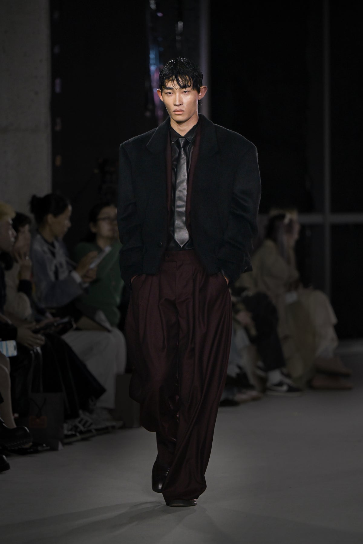 A model walks the runway wearing an oversized black blazer with exaggerated shoulders over a dark shirt and shiny gray tie, paired with loose burgundy wide-leg trousers and black shoes.