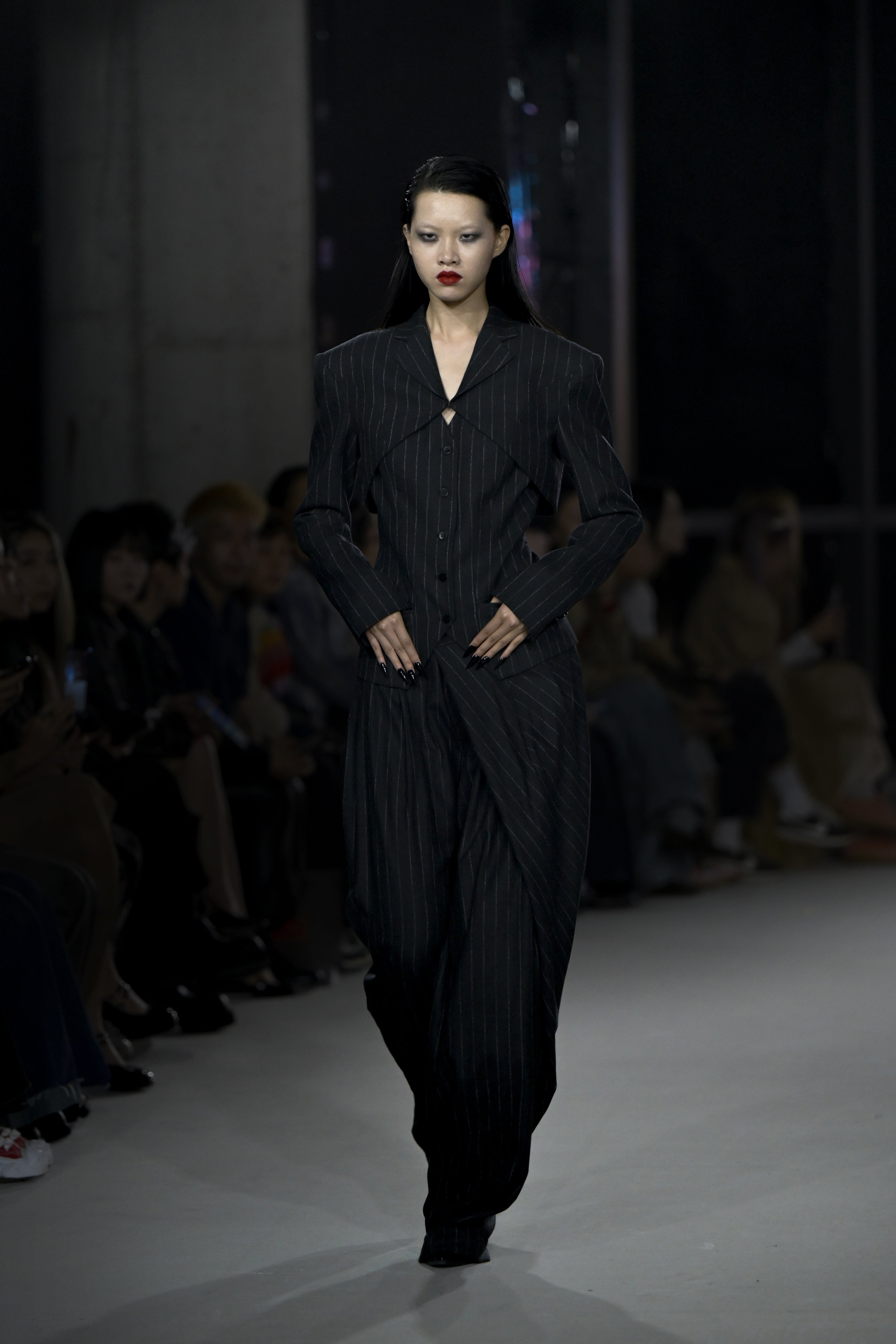 A model walks the runway wearing a dark pinstripe tailored ensemble with a sharp, angular silhouette featuring broad shoulders, a structured jacket with an open neckline, and loose-fitting trousers, embodying a modern sculptural elegance.