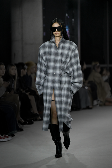 Model walking runway wearing the Oversized Flannel Shirt Dress in gradient gray plaid, featuring an elongated silhouette with a partial front opening, high collar, and voluminous sleeves paired with knee-high black boots and oversized sunglasses for a refined SHAO New York luxury bespoke tailoring look.