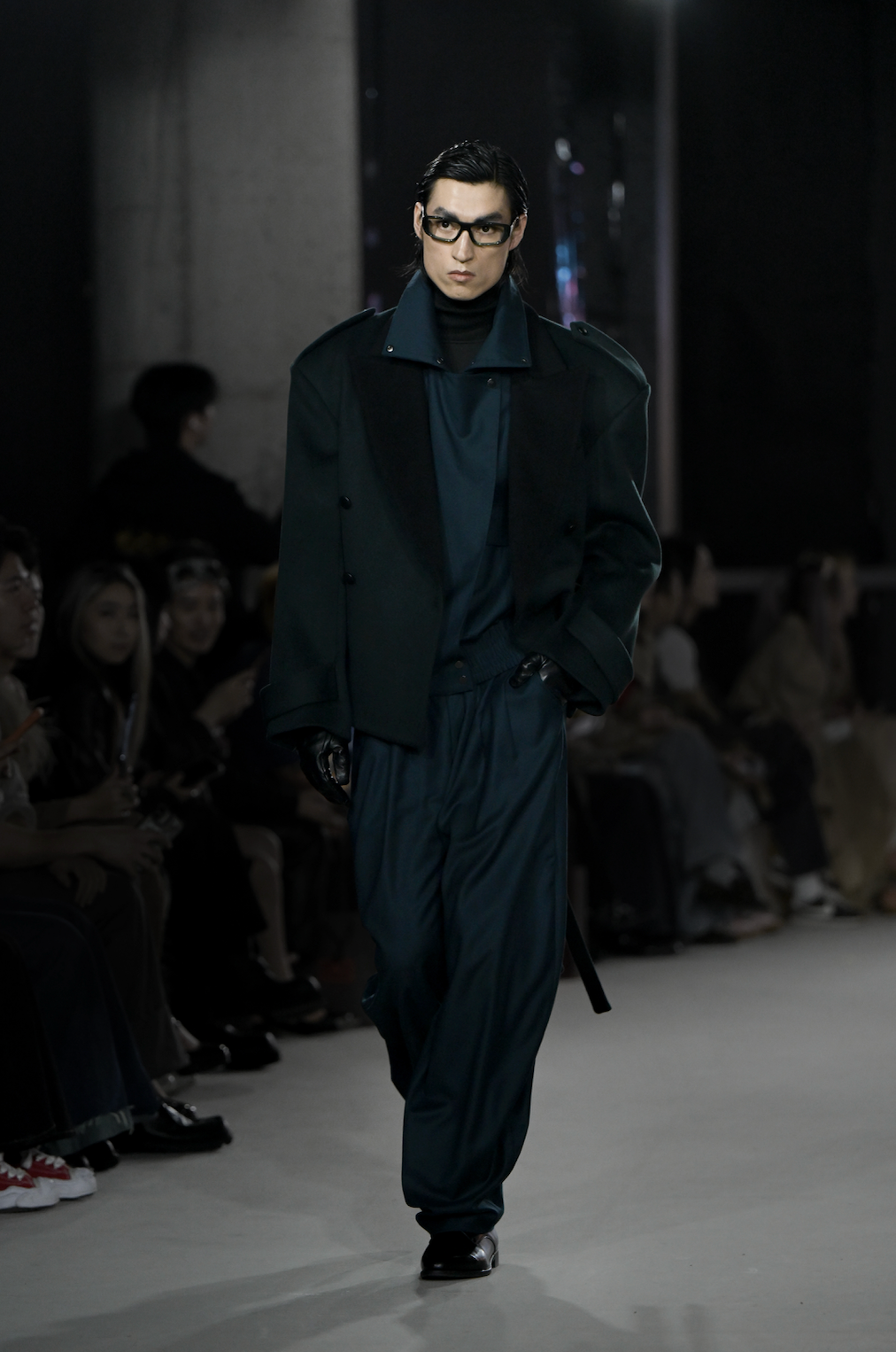 Model wearing Wool Barrel Pleated Trousers - Emerald styled with a relaxed dark tailored jacket featuring wide shoulders and a high collar, paired with black leather gloves and sleek eyewear, showcasing SHAO New York bespoke tailoring and a contemporary sculptural silhouette.