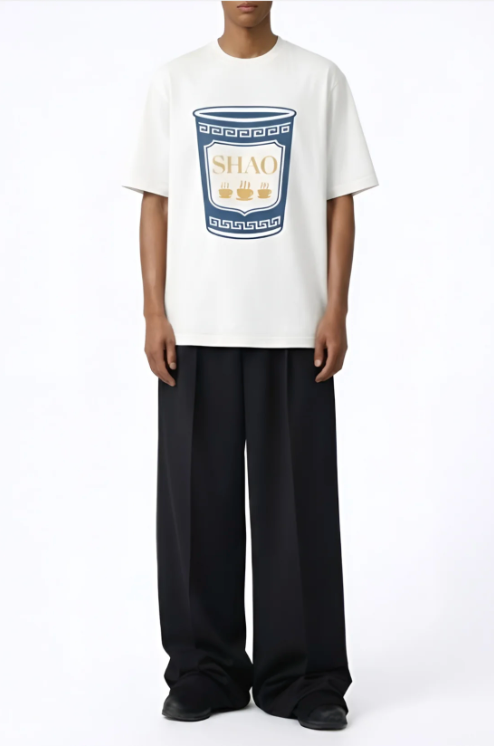 A person wearing a loose white t-shirt featuring a graphic of a blue and beige cup with the word "SHAO" and three steaming cup icons, paired with wide-leg black trousers and black shoes against a plain white background.