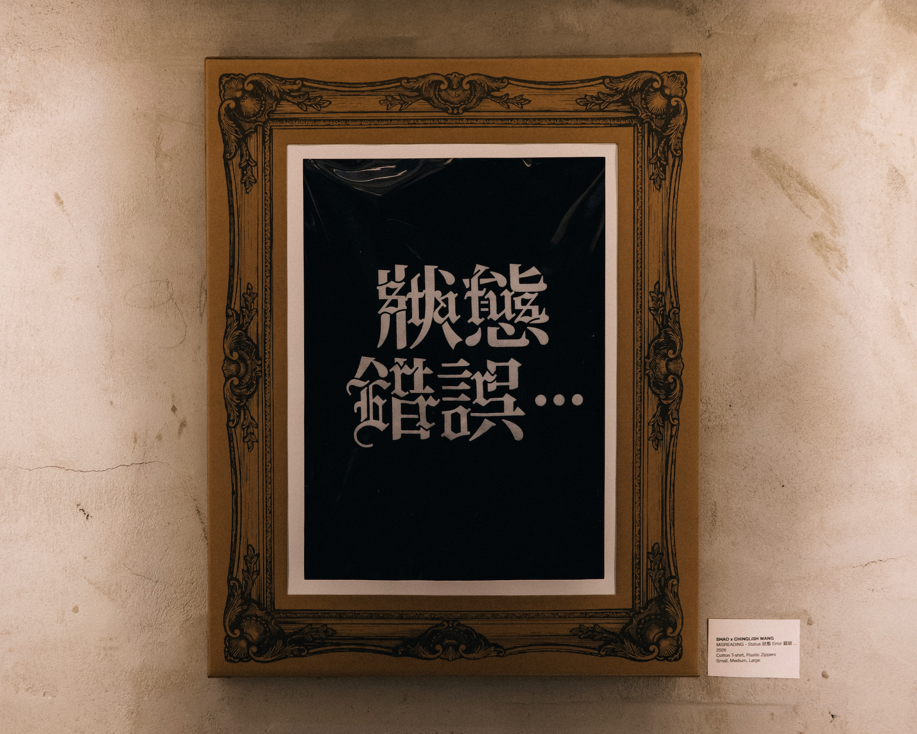 Framed artwork with ornate decorative border displaying stylized white Chinese characters on a black background, mounted on a textured beige wall with a small label nearby.