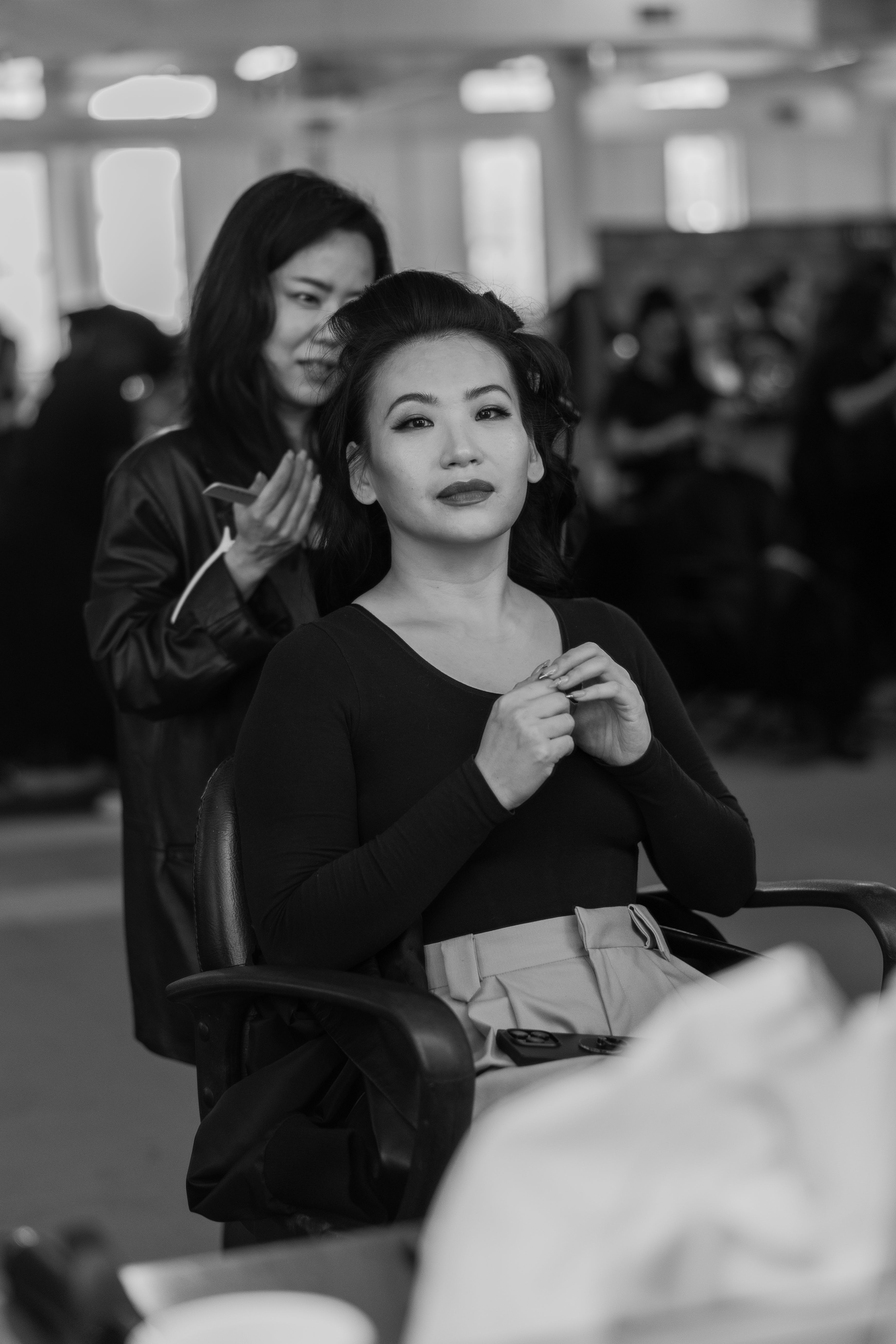 A woman with styled hair sits confidently in a chair wearing a fitted dark top and light high-waisted trousers while another person stands behind her, focusing on hair styling in a busy, softly lit room.