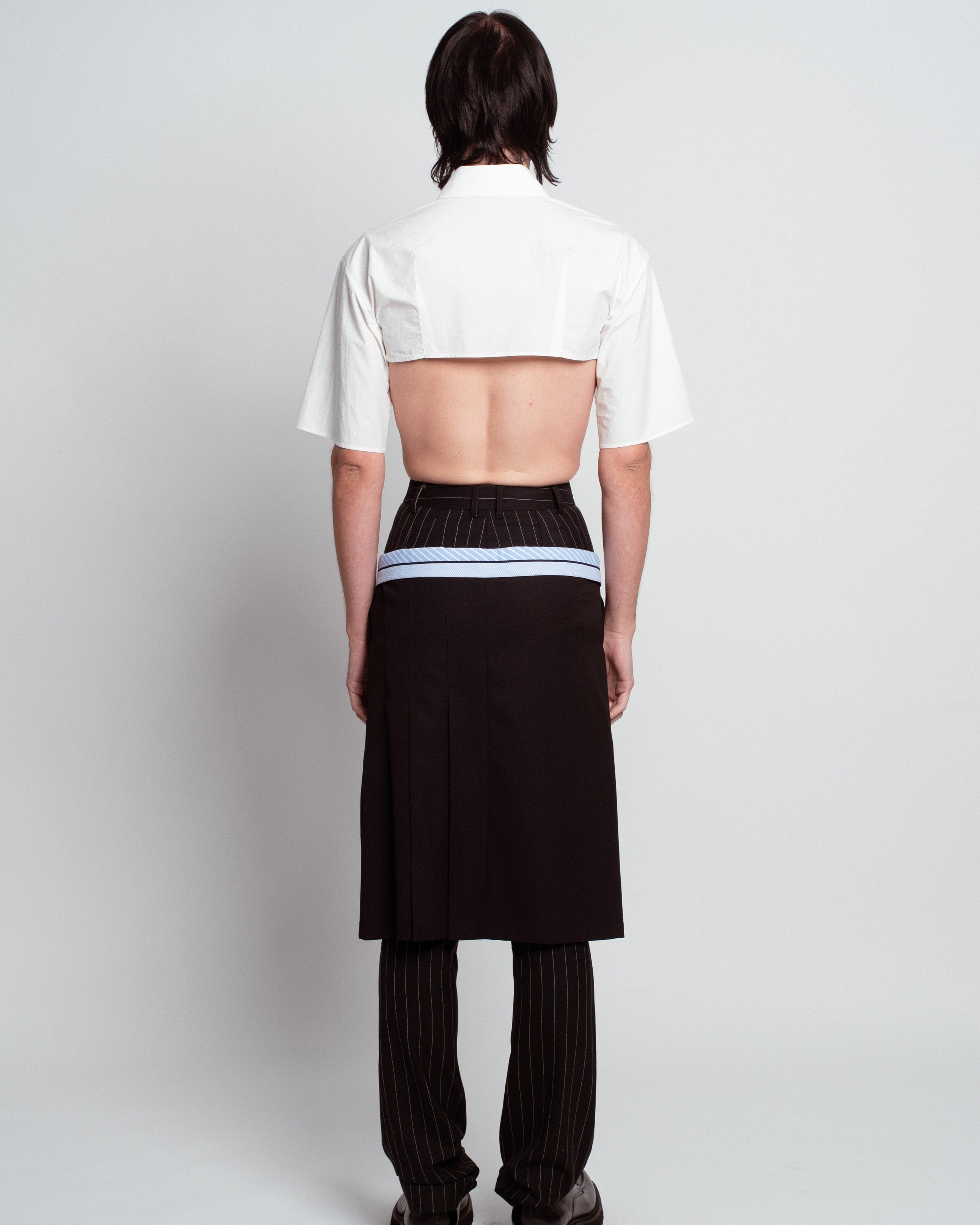 Rear view of White Cropped Utility Shirt with short sleeves and a cropped back revealing the upper back, styled with pinstriped trousers and a pleated black skirt sash for a sculptural silhouette reflecting SHAO New York bespoke tailoring and luxury designer fashion.