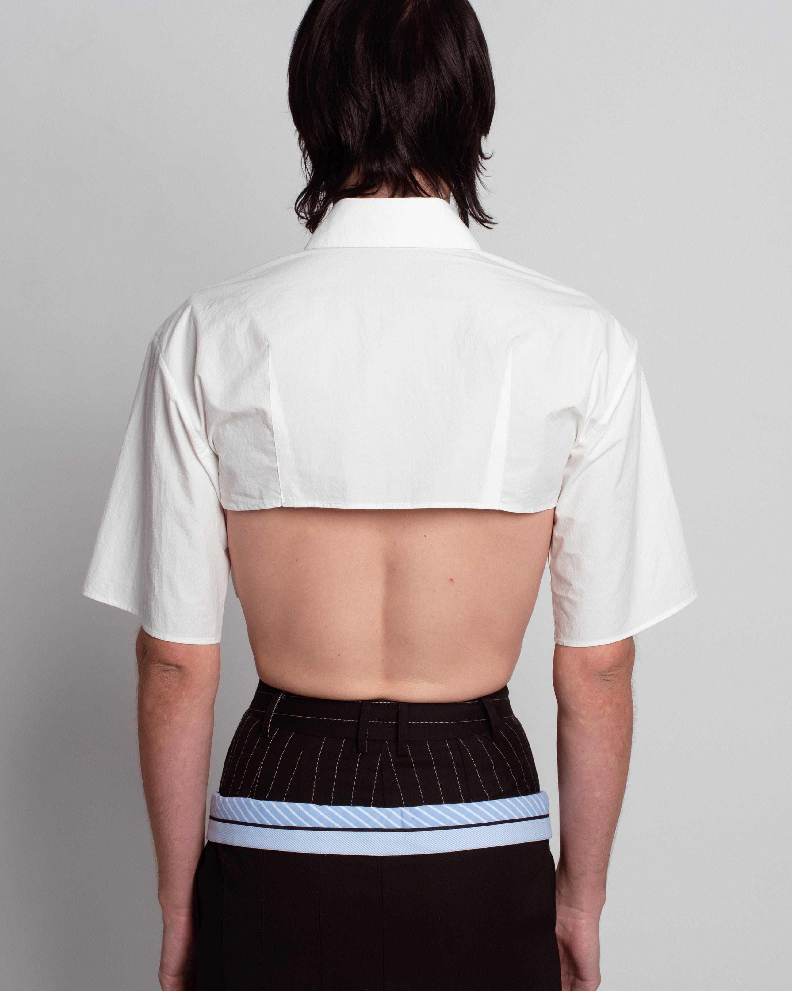 Back view of White Cropped Utility Shirt with short sleeves and a boxy cut revealing the upper back, styled with pinstriped tailored trousers and a blue belt, reflecting SHAO New York bespoke tailoring and luxury designer fashion.