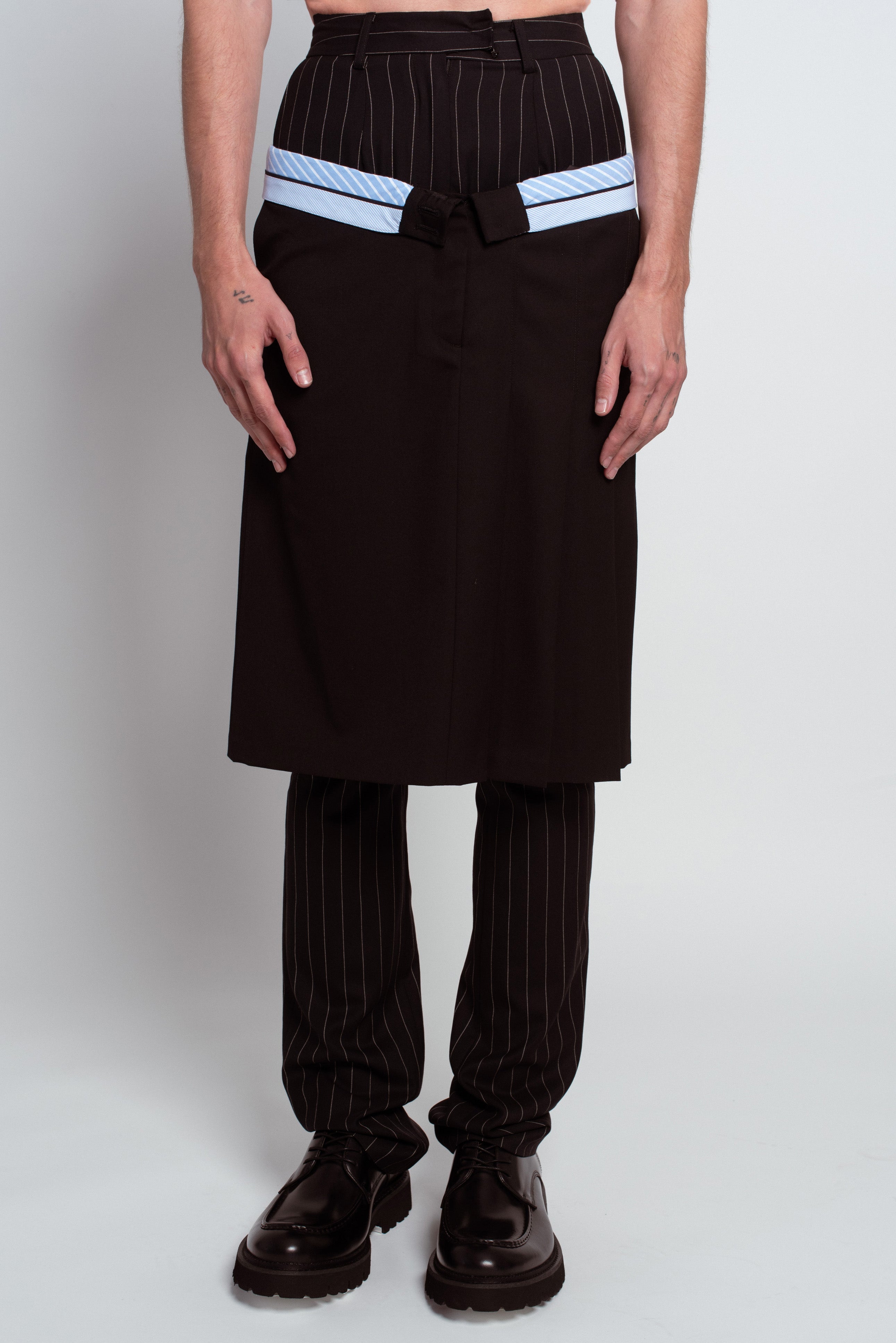 Front view of Pinstripe Layered Trousers featuring a tailored black skirt overlay with contrasting light blue striped band detail above classic black pinstriped pants, reflecting SHAO New York bespoke tailoring and luxury designer fashion with a modern sculptural silhouette.