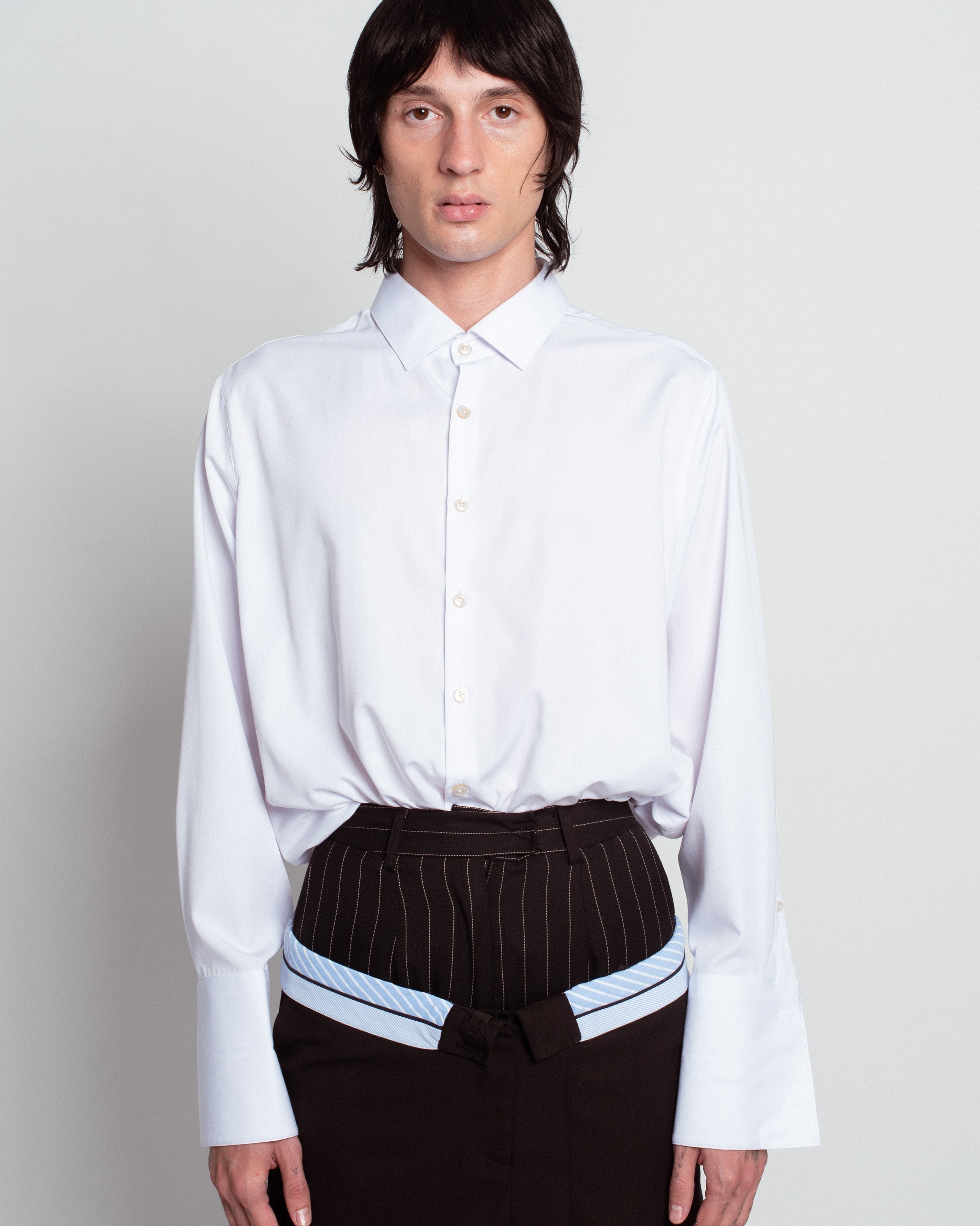 Front view of White Classic Button-Down Shirt with an oversized fit, structured collar, and elongated cuffs, styled with pinstriped tailored trousers and belt, reflecting SHAO New York bespoke tailoring and luxury designer fashion.