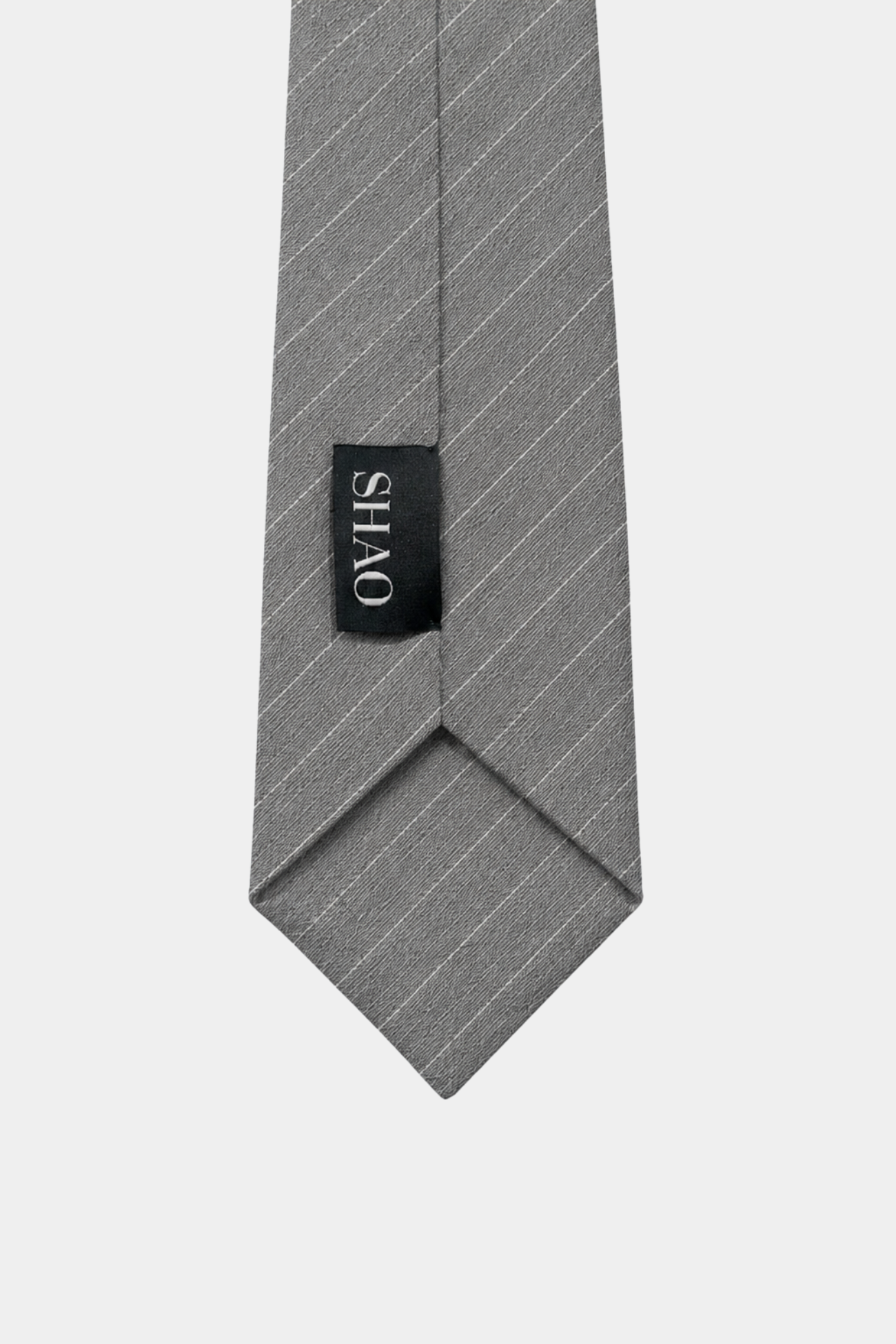 Detail of the pointed end of the Grey Pinstripe Tie 10cm showcasing fine diagonal pinstripes on a textured grey fabric and a sewn-on black SHAO label, reflecting bespoke tailoring and luxury fashion.