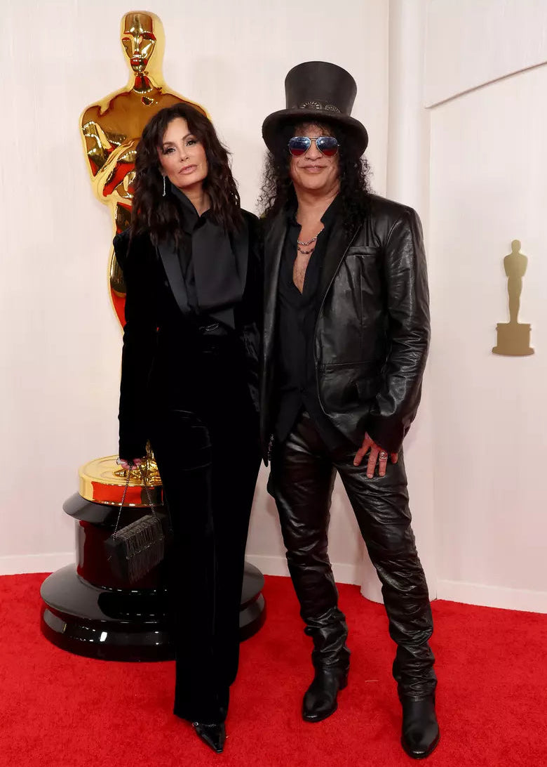 A woman in a black tailored velvet suit with a black satin blouse stands next to a man wearing a black leather jacket, leather pants, a tall top hat, and sunglasses on a red carpet with a large gold statue behind them.