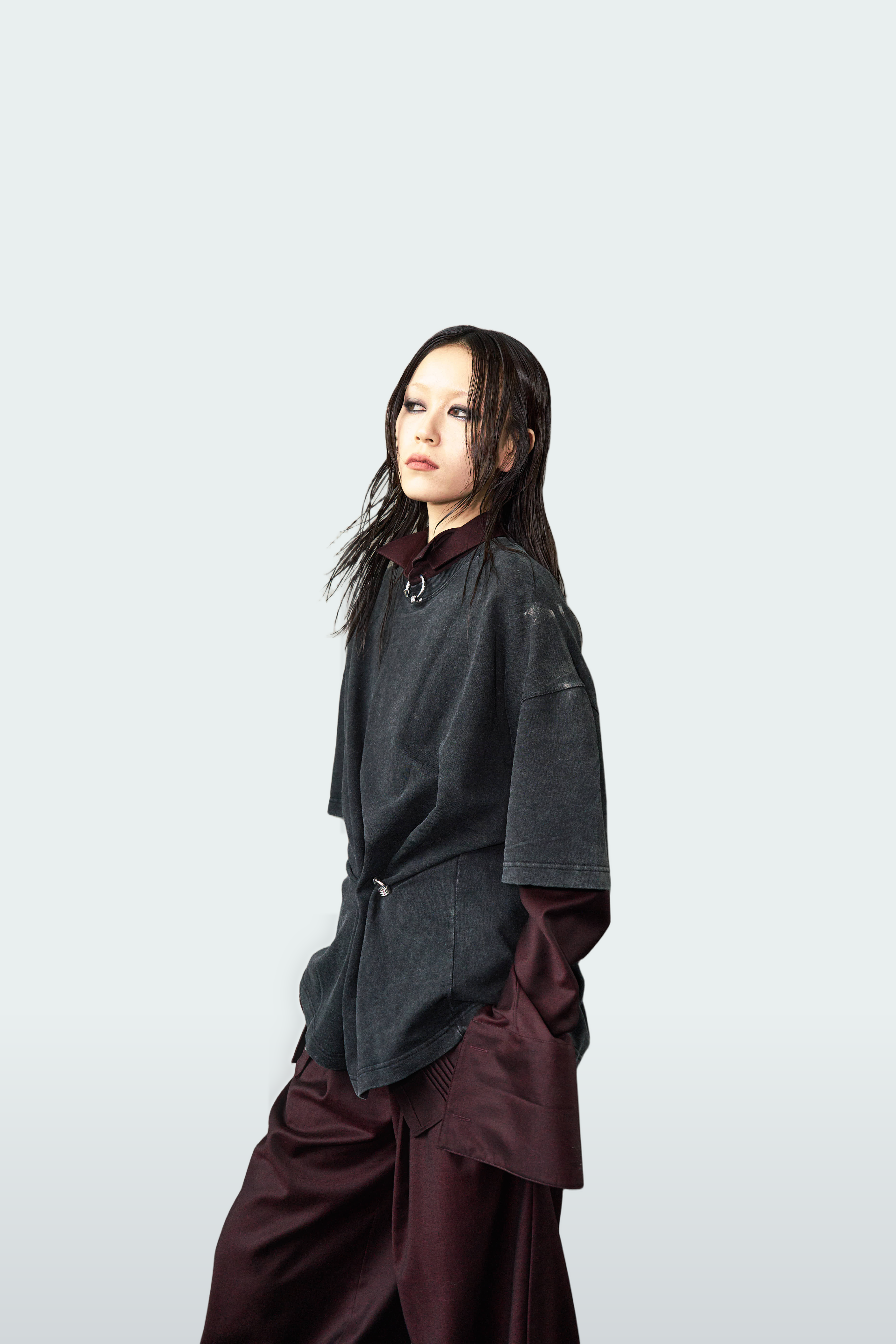 A model wearing a dark charcoal gray oversized top with a high collar and dark burgundy wide-leg pants, posed against a solid light gray background.