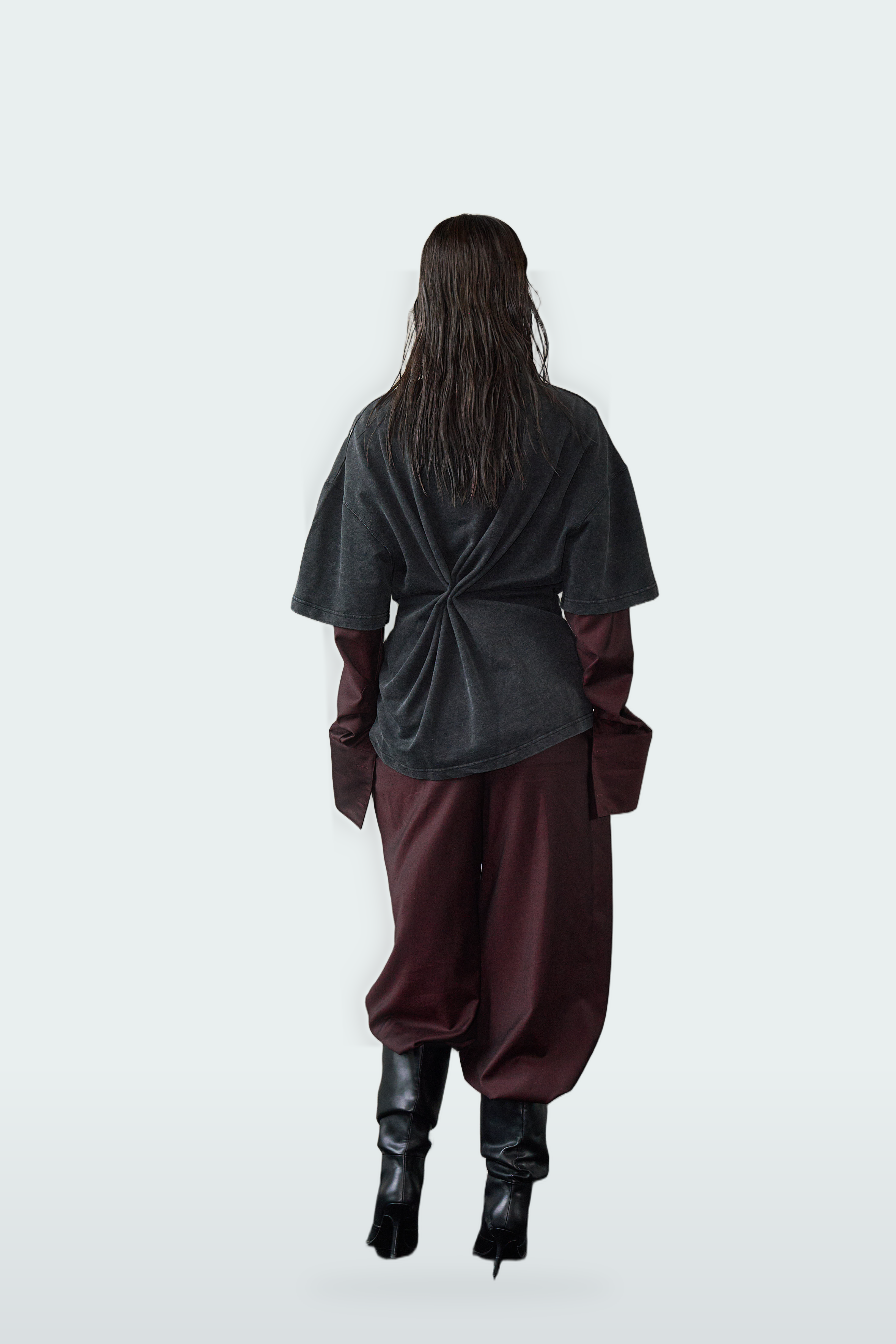 A model viewed from behind wearing a dark gray oversized top with a twisted back detail, maroon baggy pants, and black high-heeled boots against a light gray background.