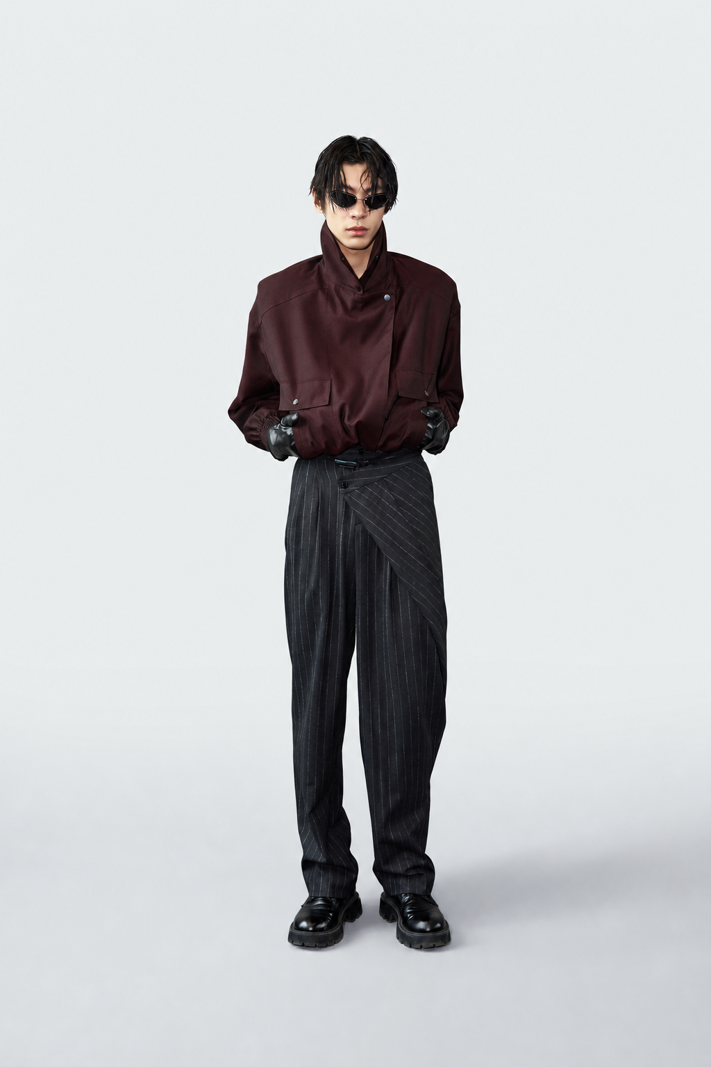 A model stands against a light gray studio background wearing a dark burgundy high-collar shirt, black pinstriped trousers, black gloves, and black shoes.