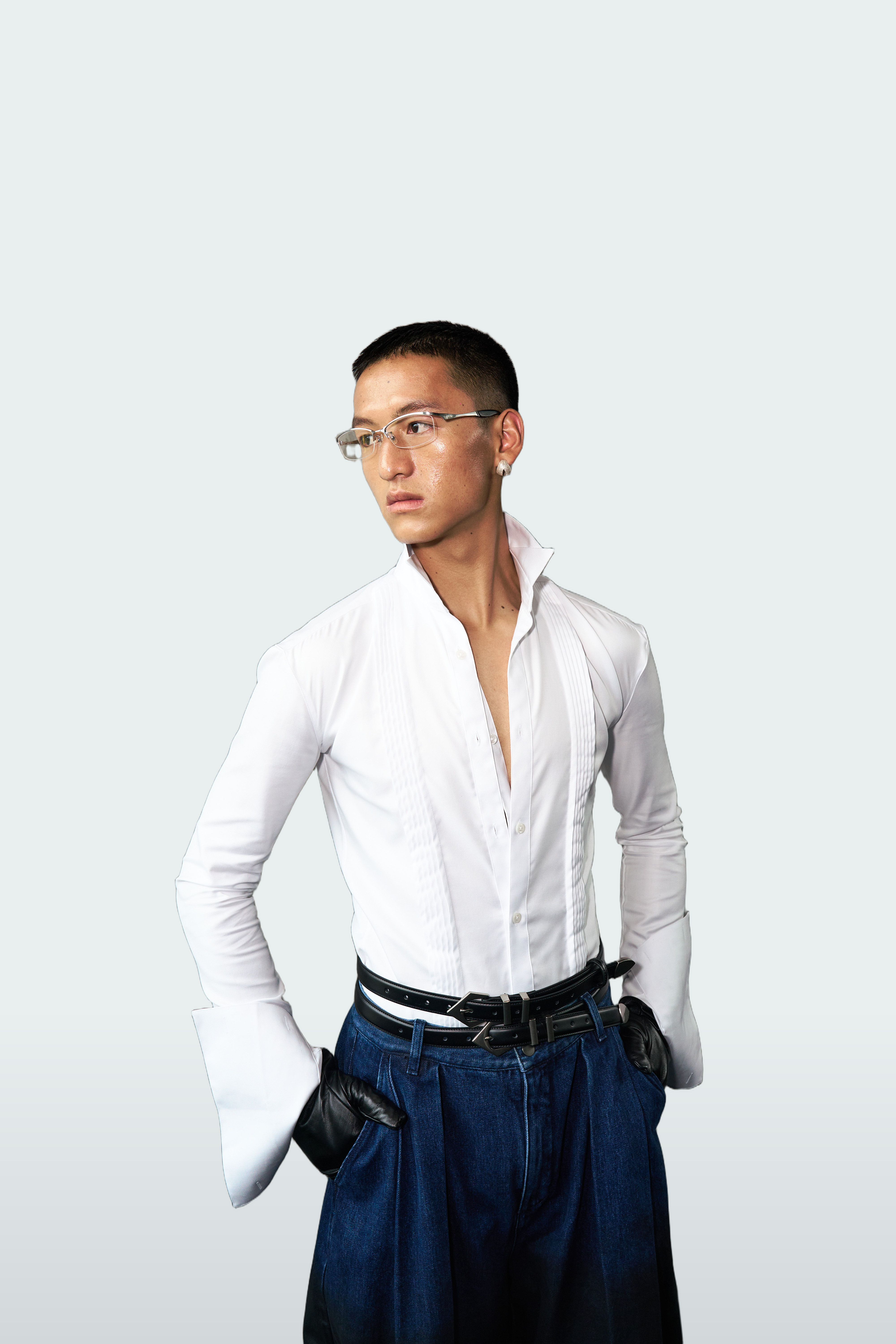 A man wearing a white long-sleeve button-up shirt, dark blue jeans, a black belt, and black gloves, standing against a light gray studio background.