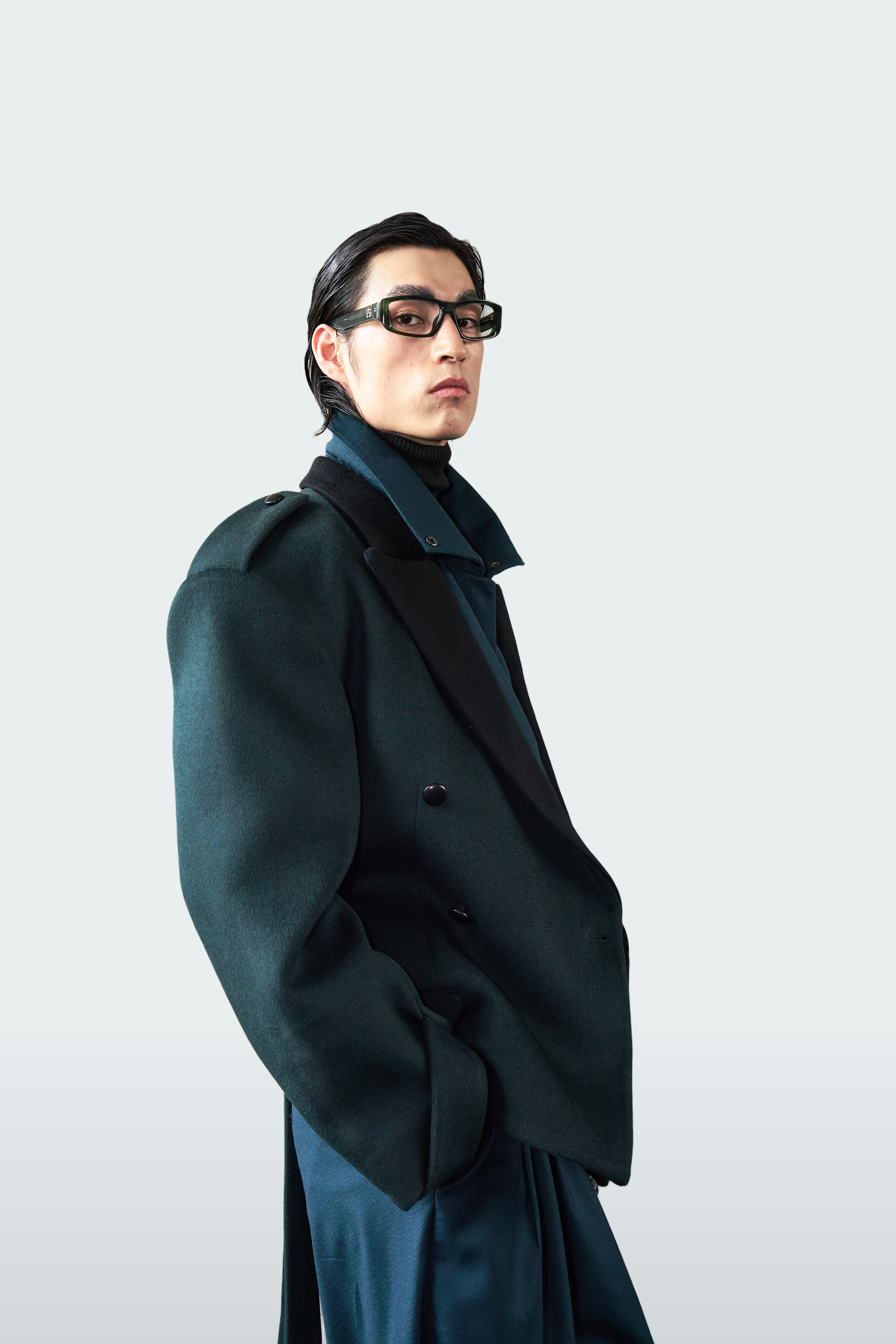 A model wearing a dark teal double-breasted overcoat over a matching dark turtleneck and trousers, styled with black rectangular eyeglasses against a light gray studio background.