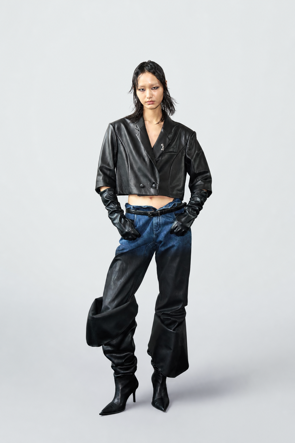 A model stands against a light gray studio background wearing a black cropped leather jacket, blue jeans, black gloves, and black heeled ankle boots.